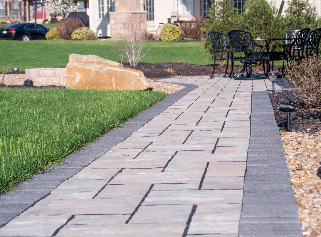 Stone walkway bordered by dark pavers, leading to patio with black furniture.
