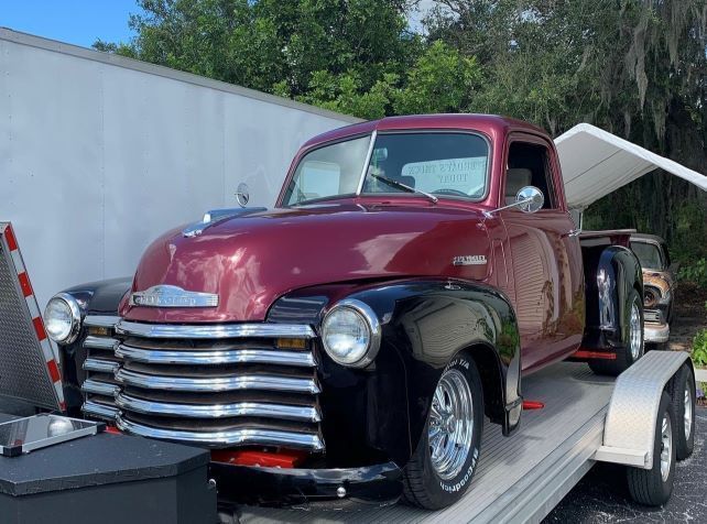 Maroon vintage pickup truck on a trailer, parked outdoors near a white building