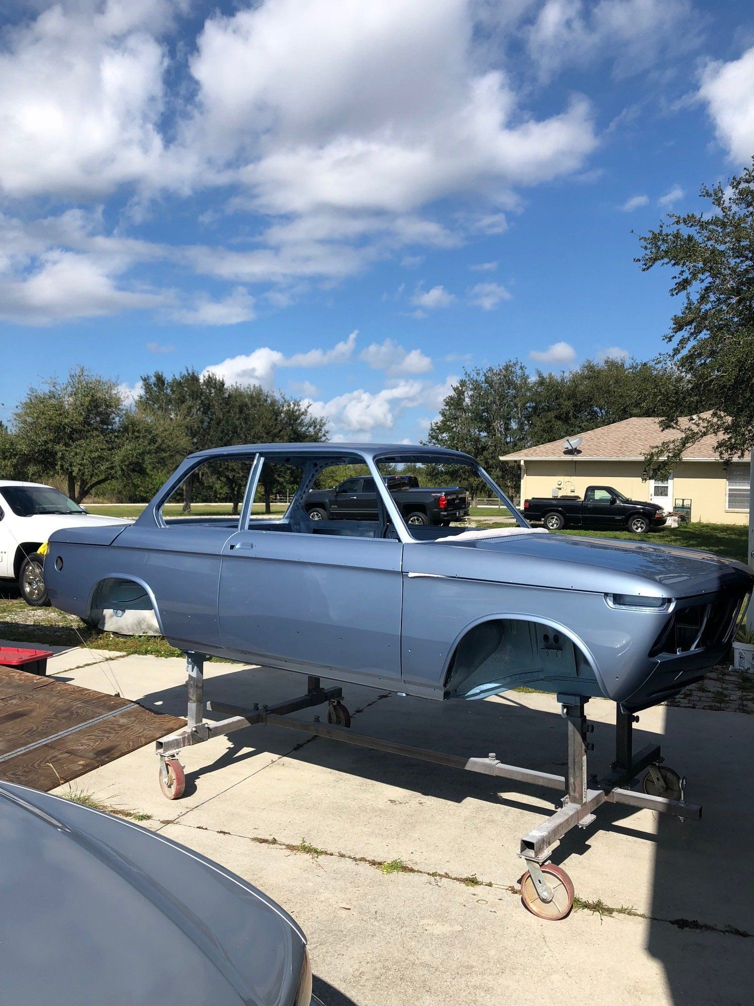 Blue vintage car body on a dolly in a driveway, with trees and houses in the background.