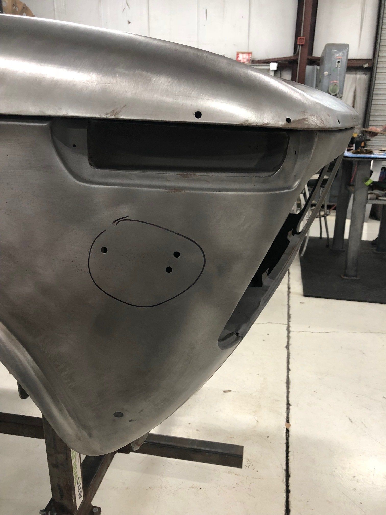 Close-up of a gray metal vehicle body panel with a black opening and a marked oval area, in a workshop.