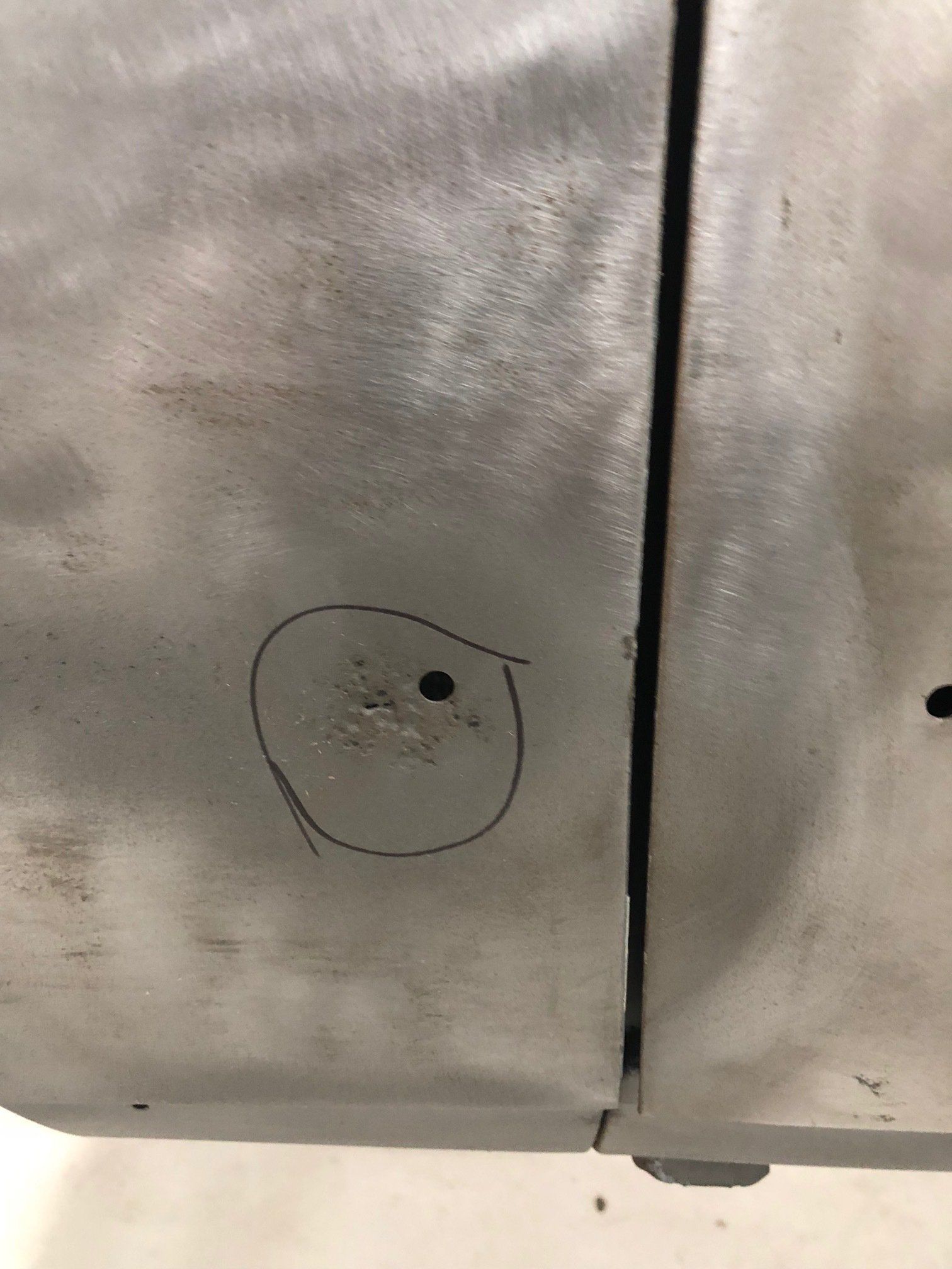 Close-up of a scratched gray metal surface with a small circled hole and black dot near a seam.