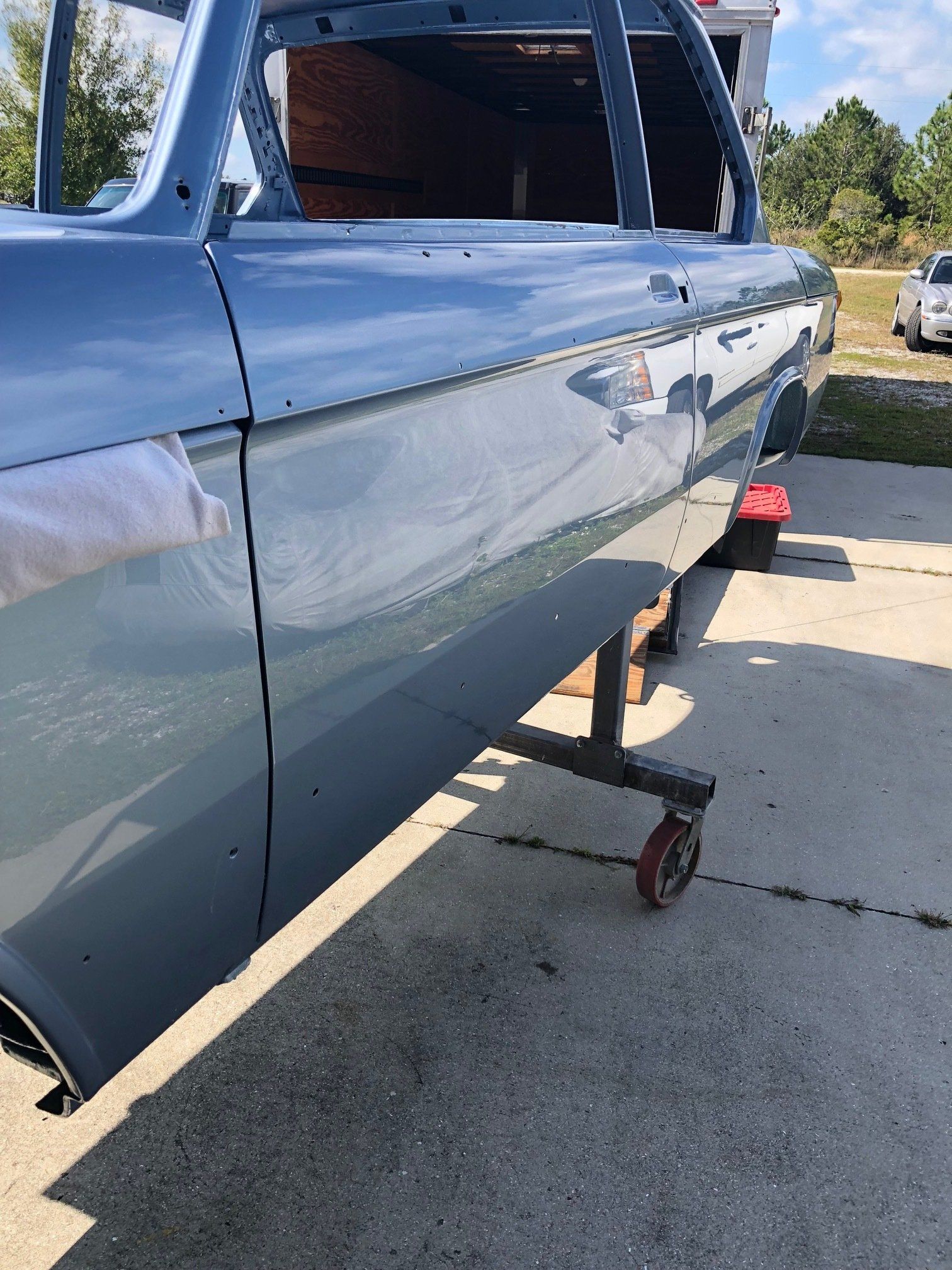 Blue car body on a dolly in a driveway, with a rear window opening and red tape on the side.