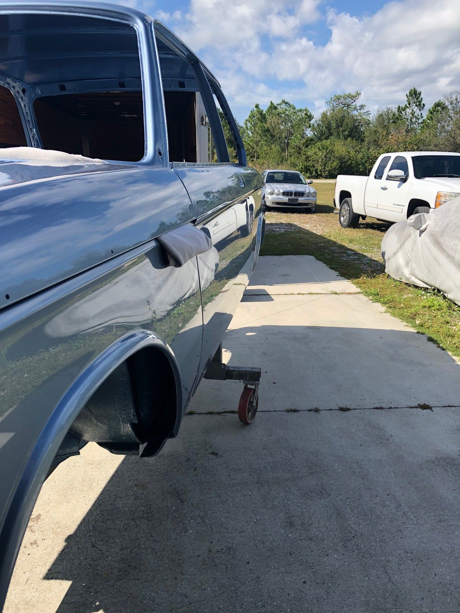 Blue car body on jacks in a driveway, with other vehicles parked nearby in bright sunlight