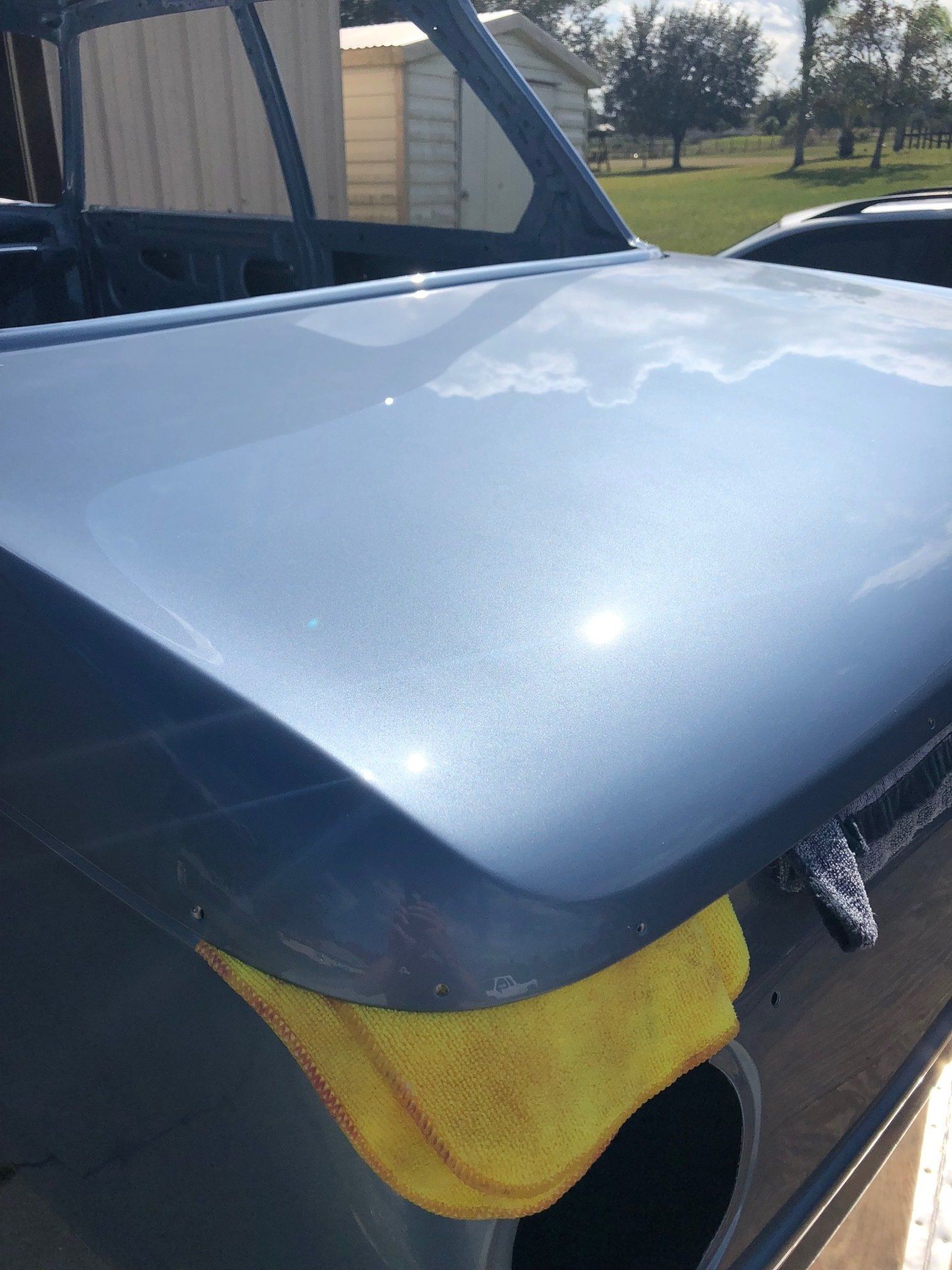 Blue car hood propped open with yellow cloth underneath, outdoors in daylight.