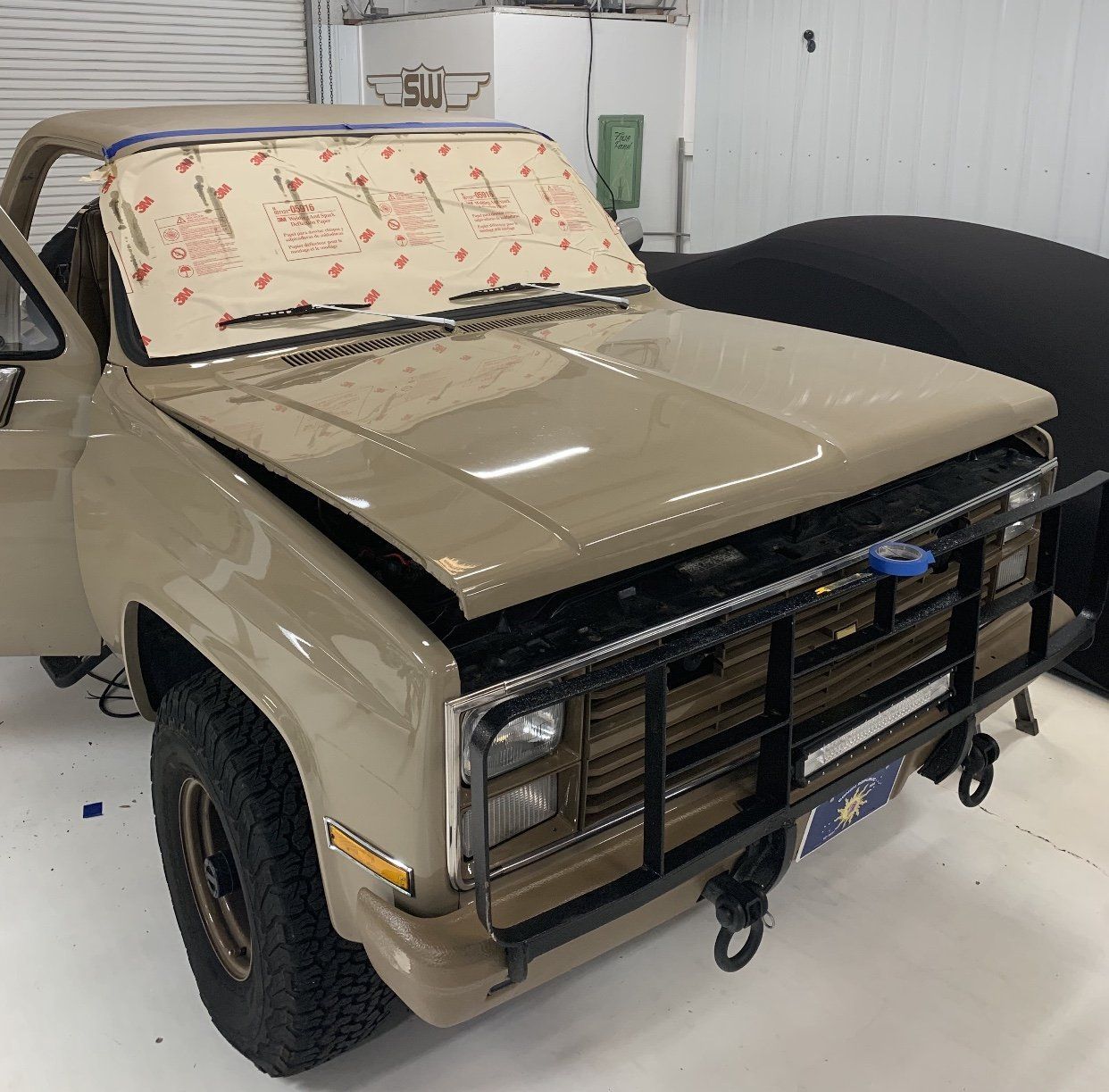 Tan military-style pickup truck with a splattered windshield and heavy front grille in a garage