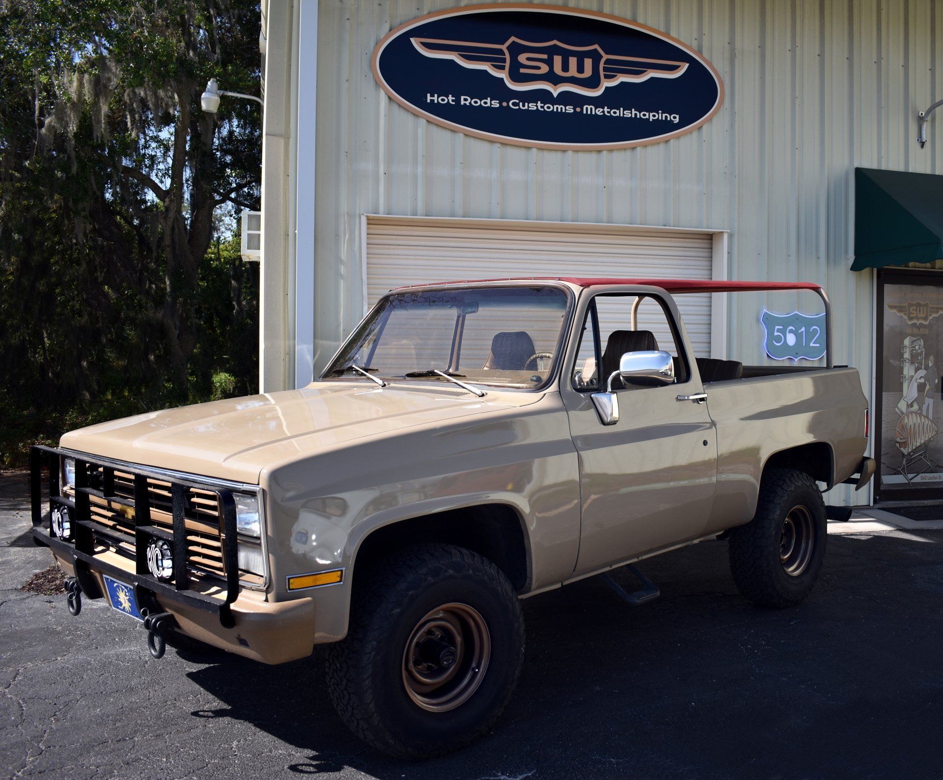 Beige vintage pickup truck parked outside a shop with a winged sign above it.