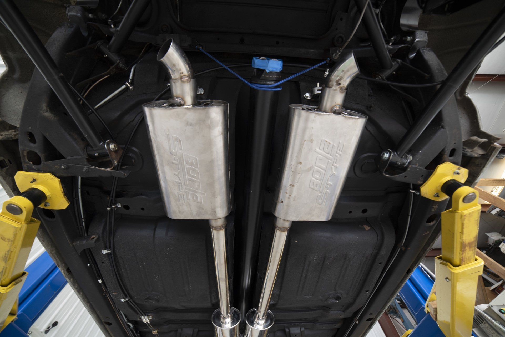 Underside of a vehicle on a lift, with dual silver exhaust mufflers and yellow support arms visible.