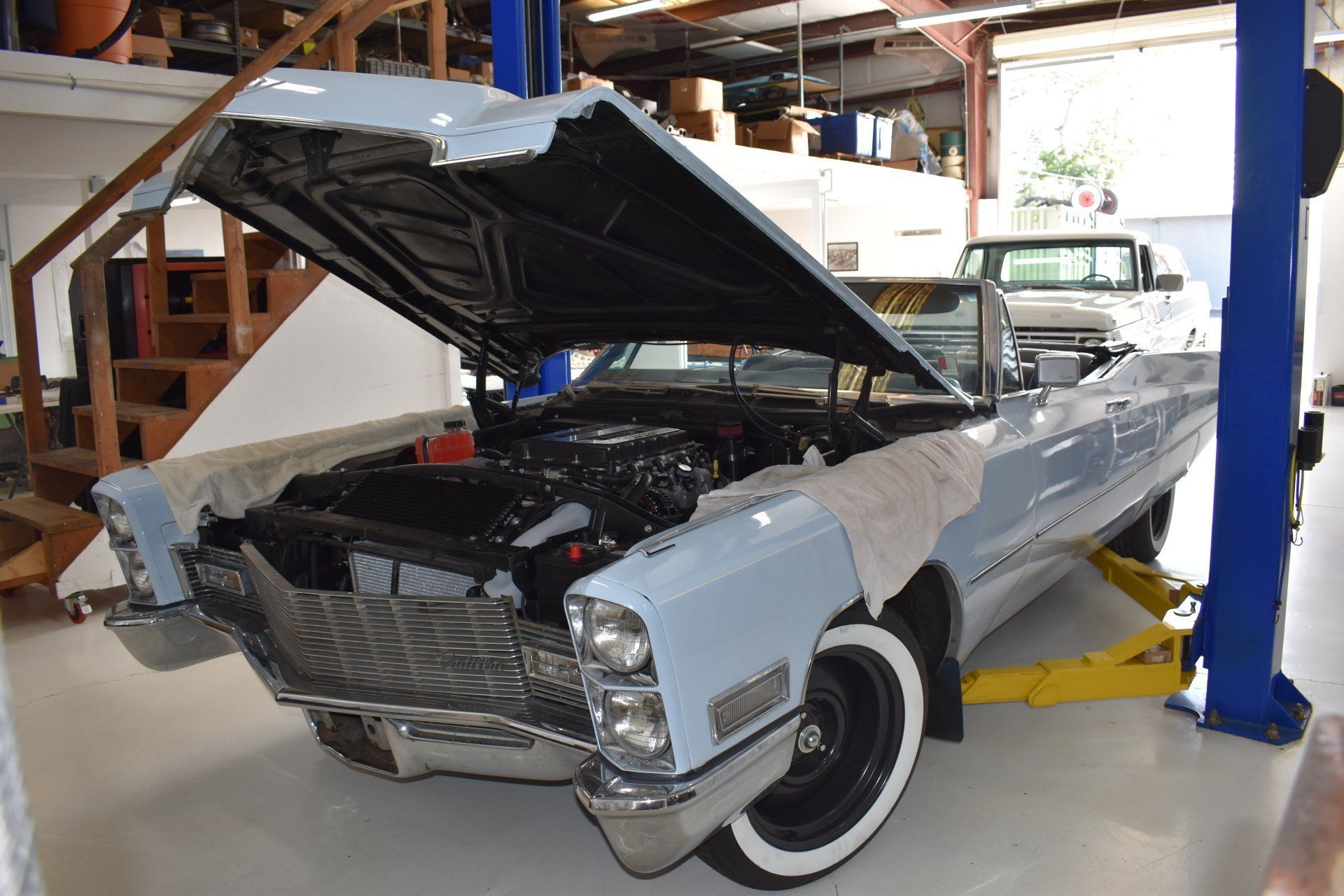 Classic white convertible in a garage with its hood open, showing the engine; another car is visible behind it.