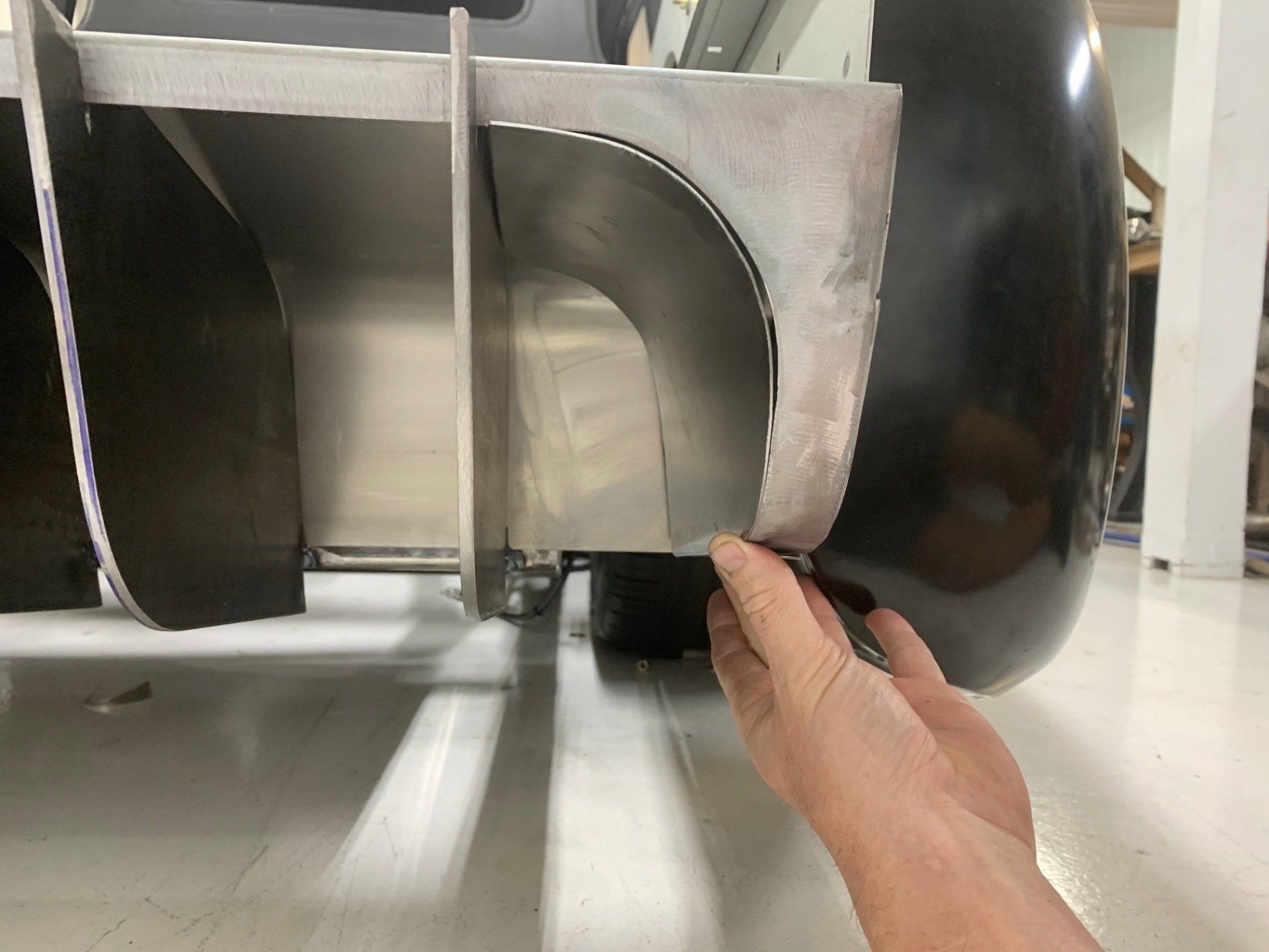 Hand reaching under a metal appliance on a white floor, near a black wheel and curved metal housing