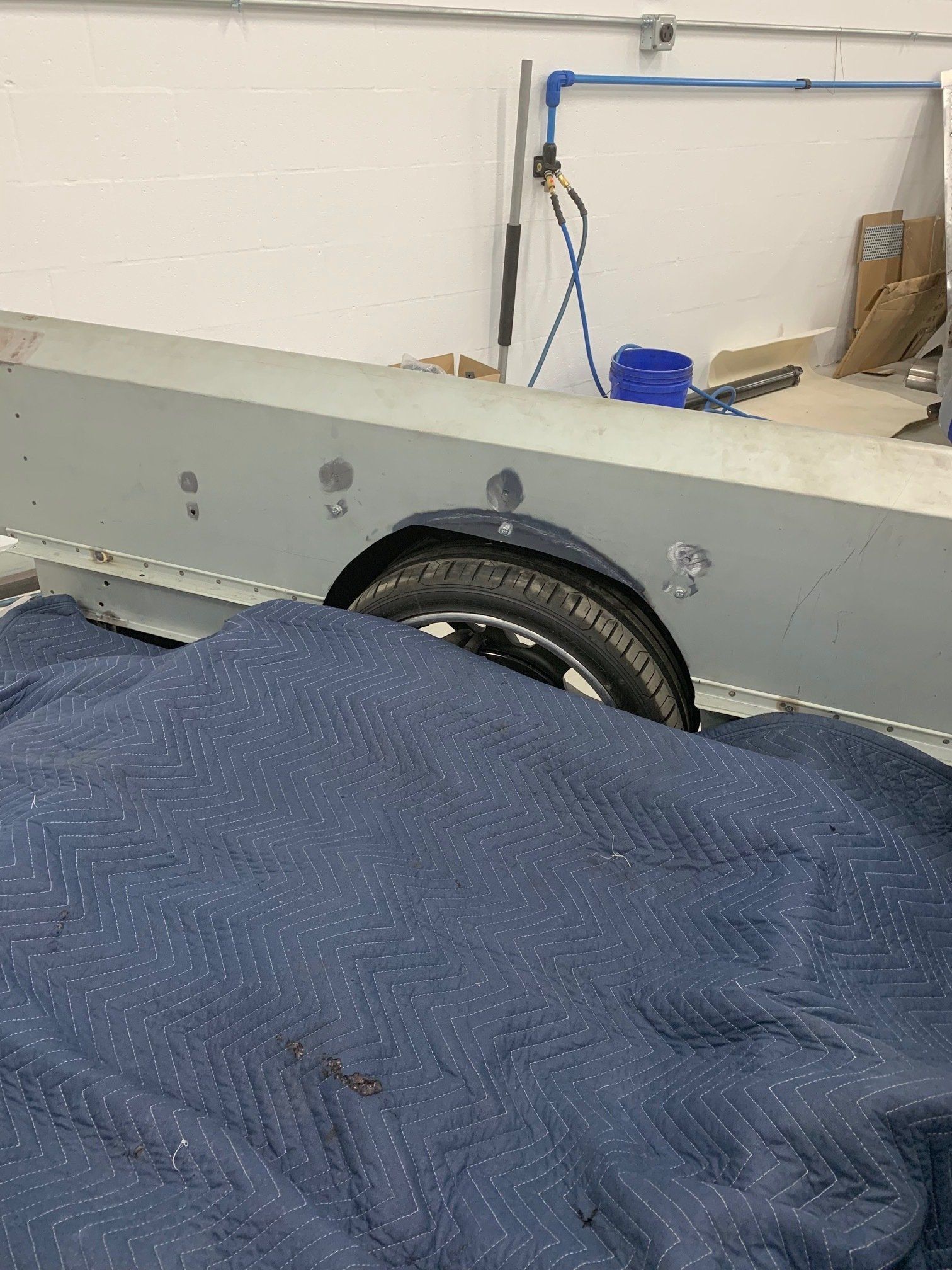 Gray vehicle body in a workshop, with a visible wheel and a blue tarp covering the foreground.