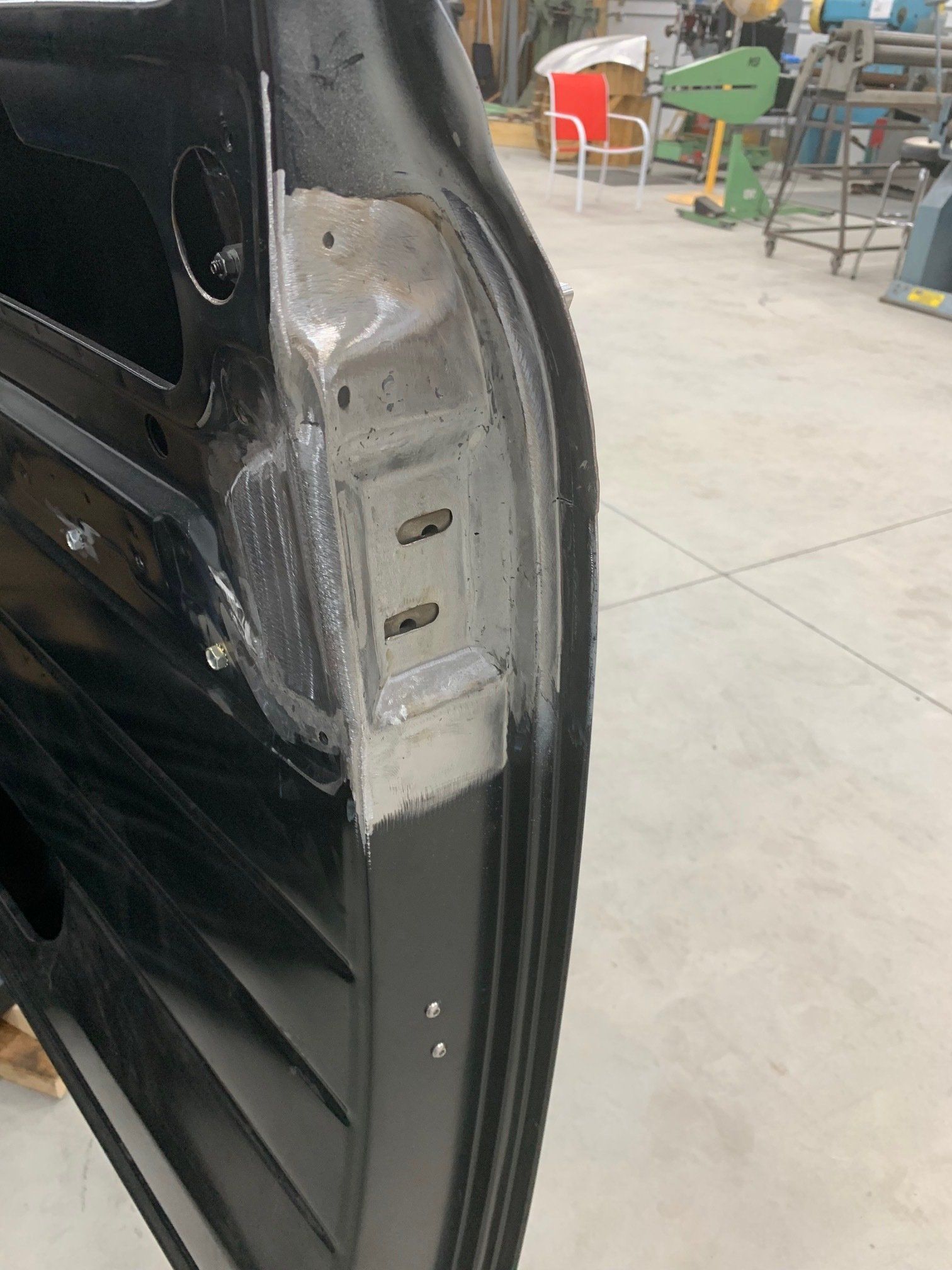 Close-up of a black car door hinge and latch area with exposed metal in a garage.