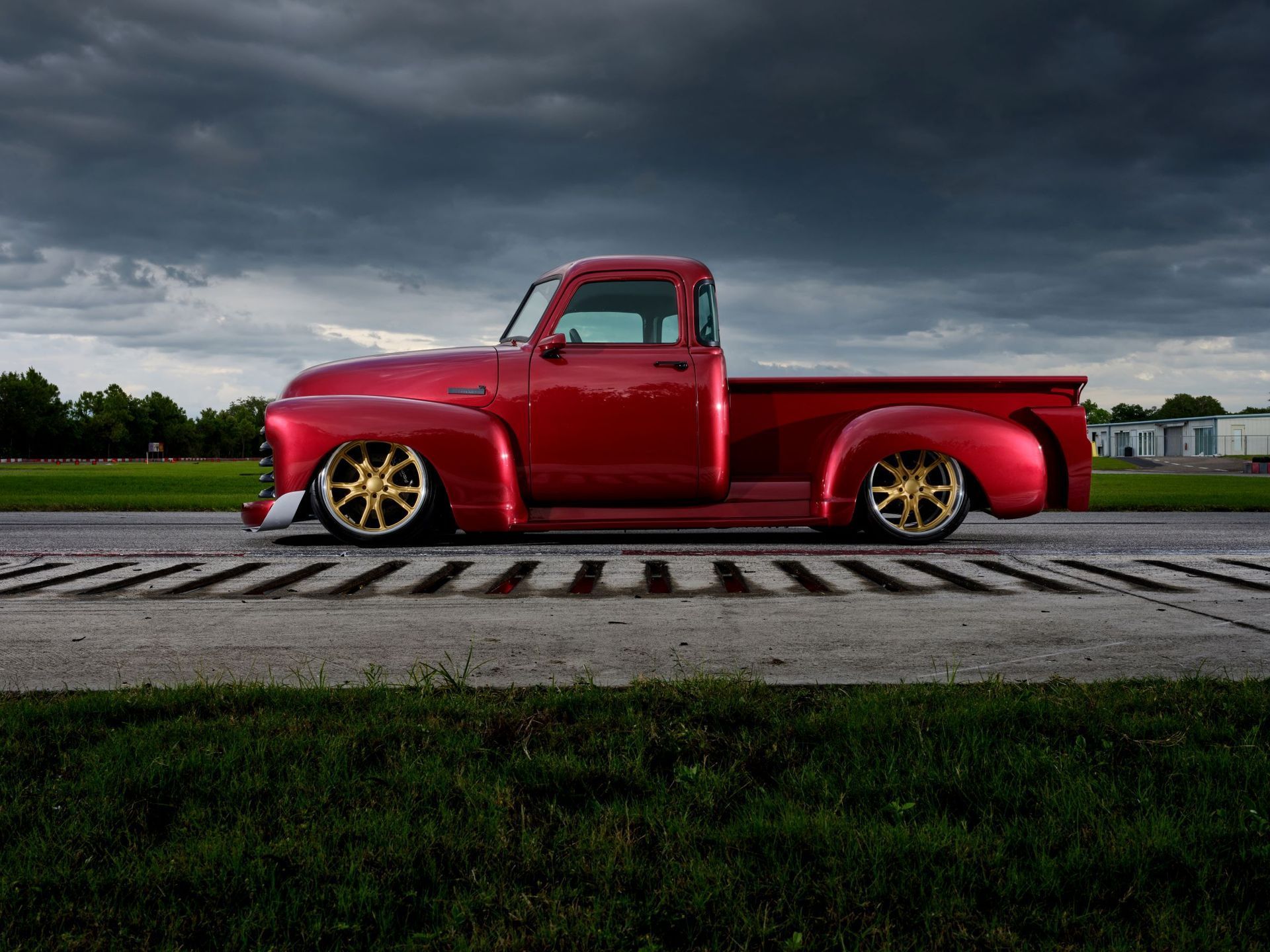 Red vintage pickup truck with gold rims parked on a road under dark storm clouds