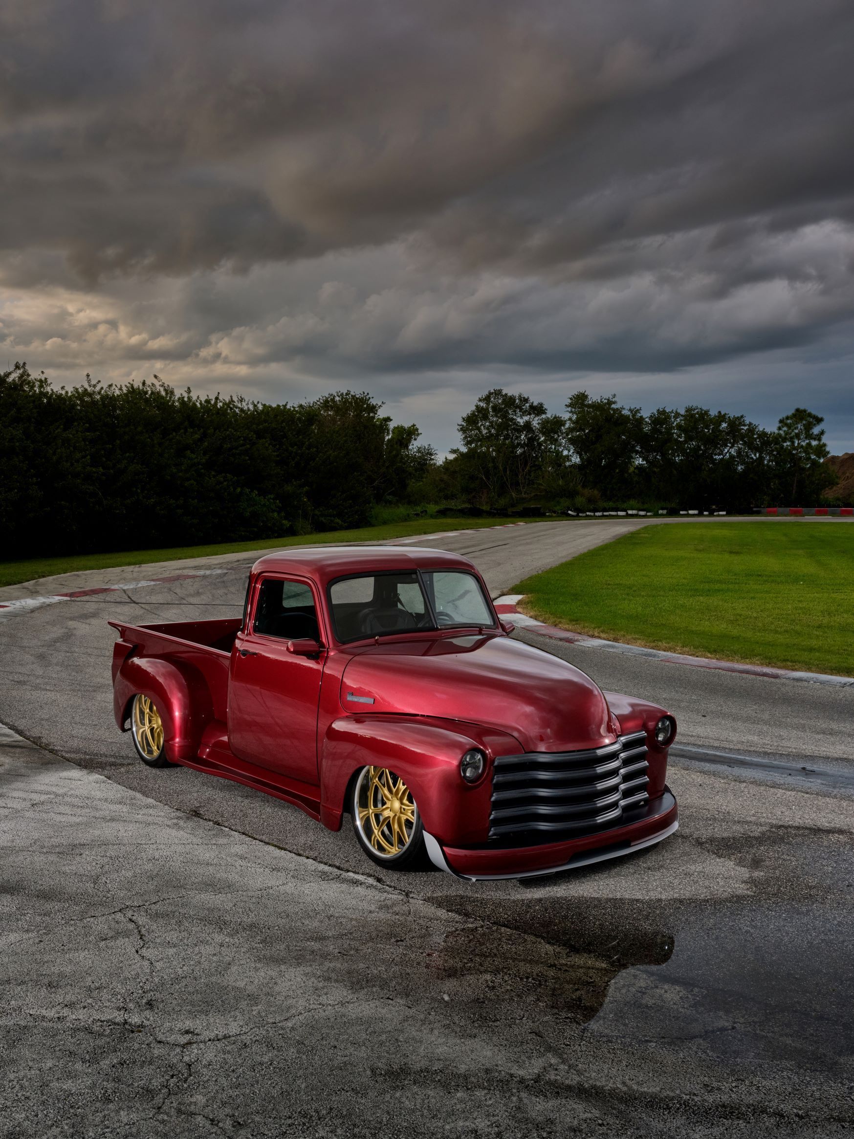 Red vintage pickup truck on a gravel lot under dark storm clouds.