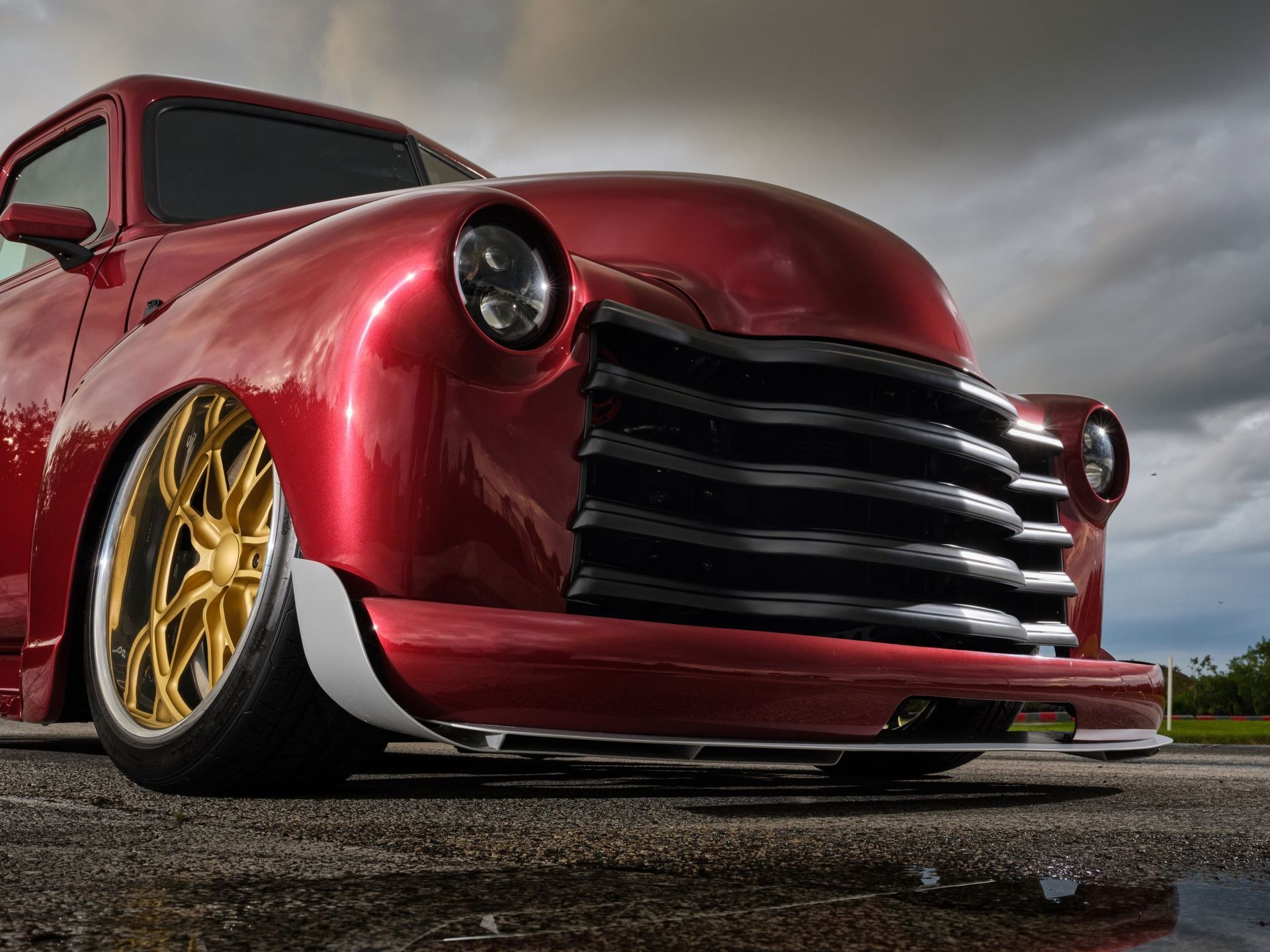 Low red custom hot rod with gold rims parked on wet pavement under cloudy sky