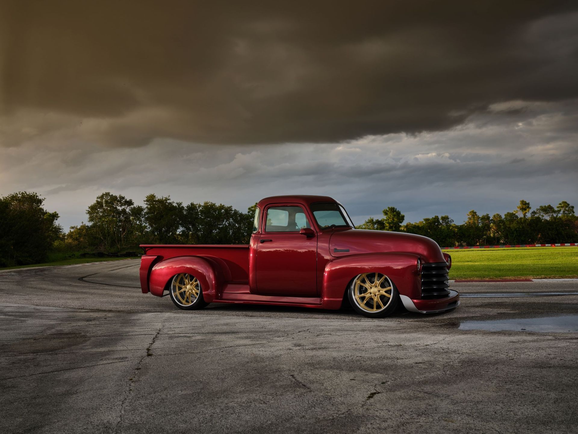 Red vintage pickup truck parked on pavement under a dark cloudy sky, side view.