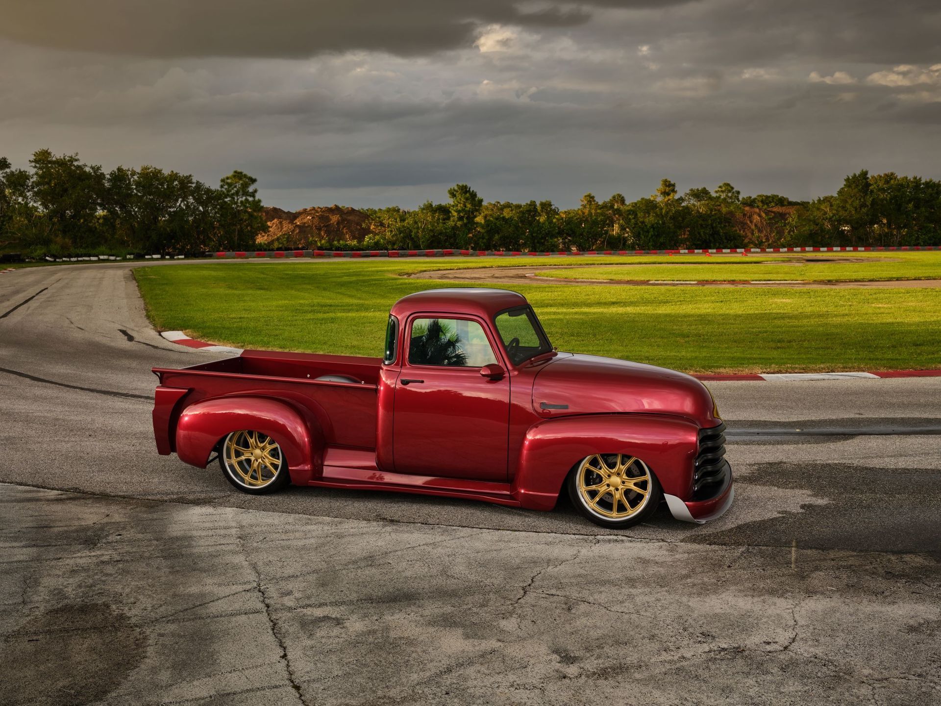 Red custom pickup truck with gold rims parked on a paved track under cloudy skies.