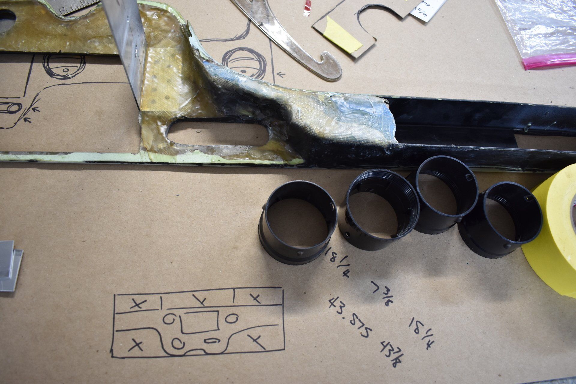 A workbench with a long dark tool, four black rings, a yellow tape roll, and sketch notes on cardboard