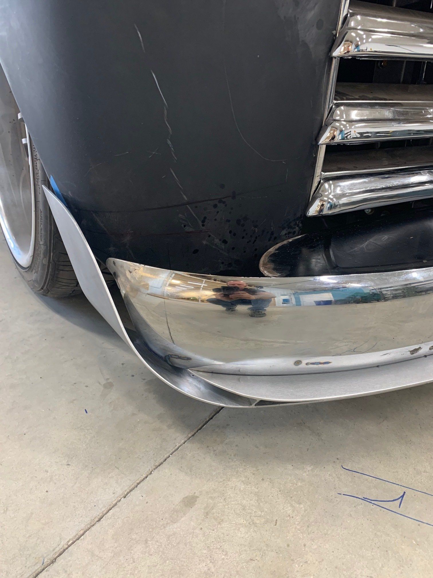 Close-up of a black car’s front bumper with chrome trim, showing scuffs and a dent near the curb.