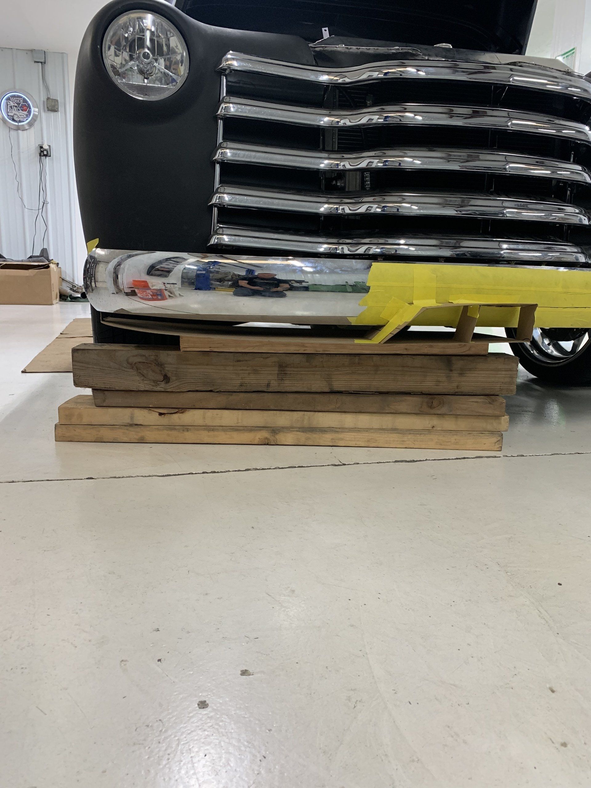 Front of a black vintage truck resting on wooden blocks, with yellow tape across the chrome bumper.