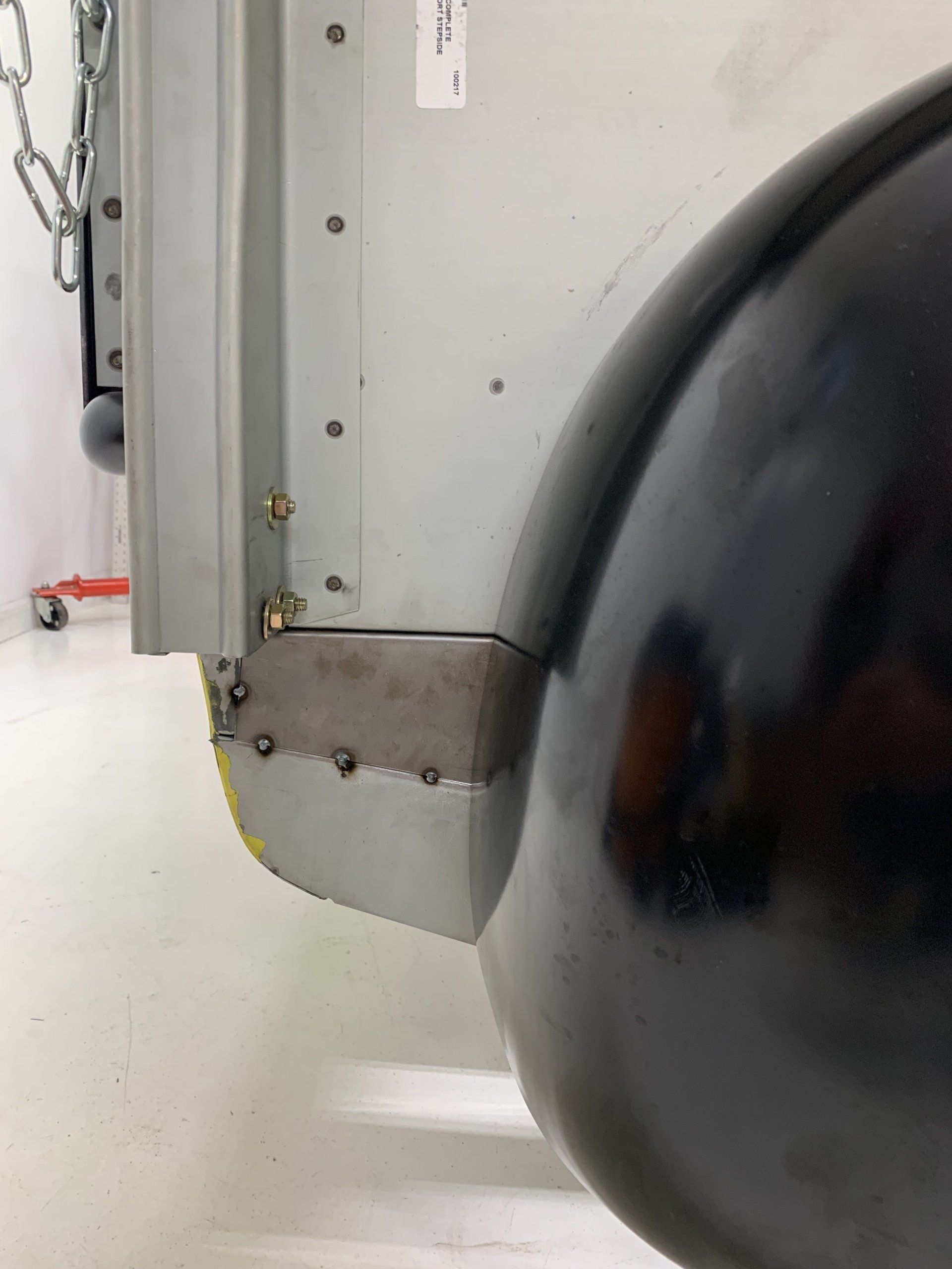 Close-up of a black wheel beside a gray metal panel with bolts and a chain on a white wall