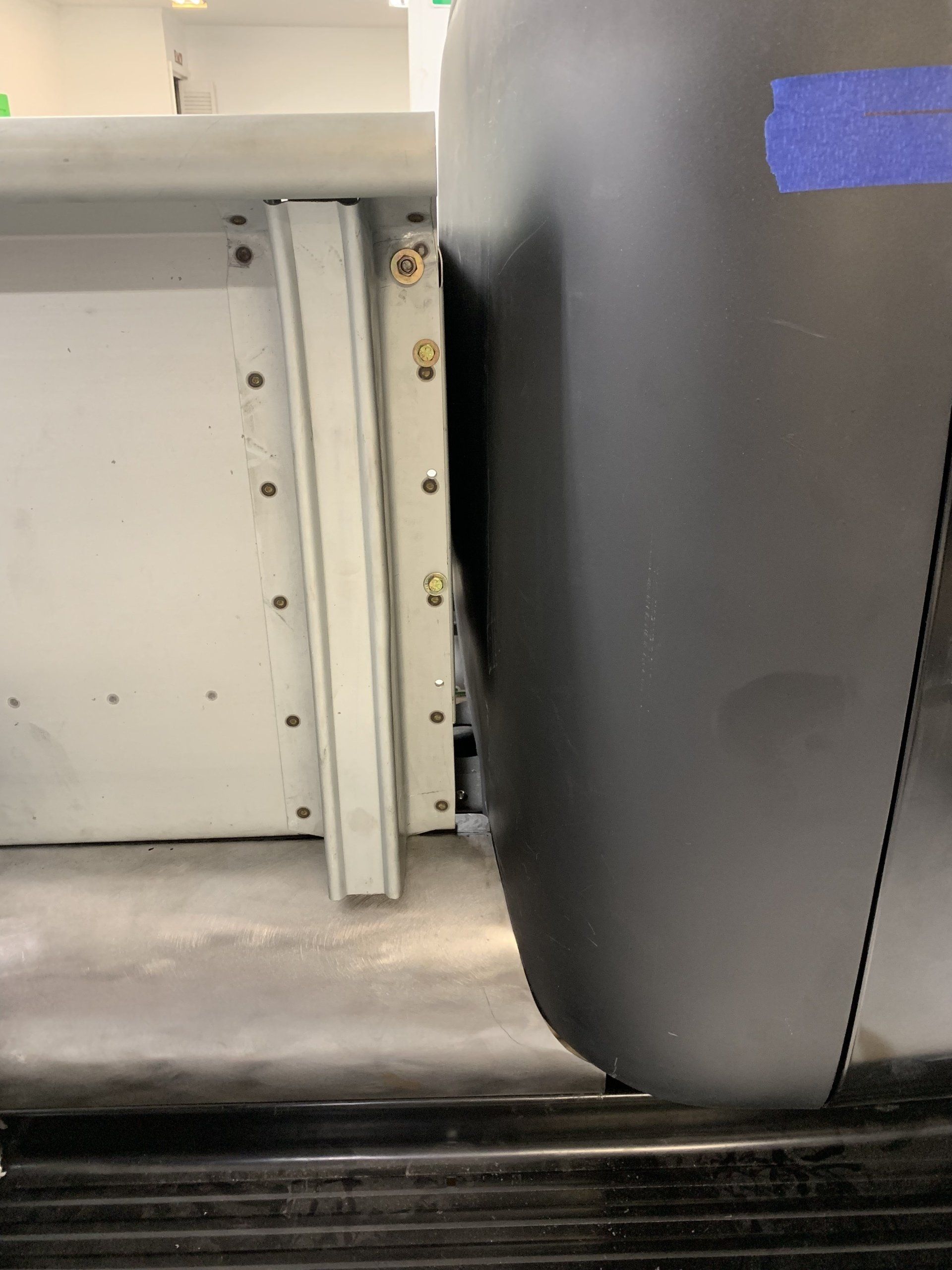 Close-up of a white cabinet hinge beside a dark gray seat with blue tape.