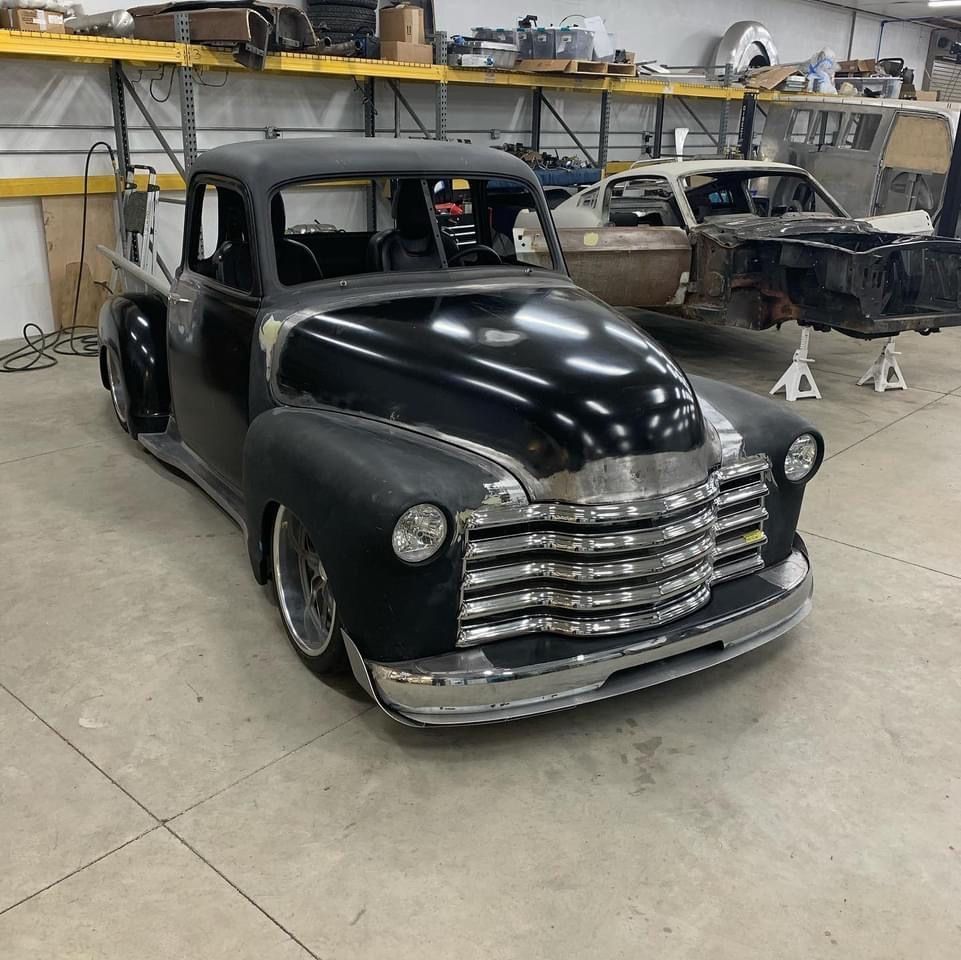Black vintage pickup truck with chrome grille in an auto shop, with a partially disassembled car behind it