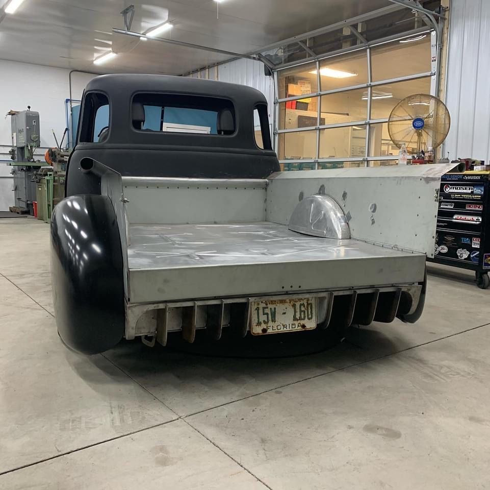 Vintage black pickup truck in a workshop, viewed from the rear with a flatbed and exposed tailgate.