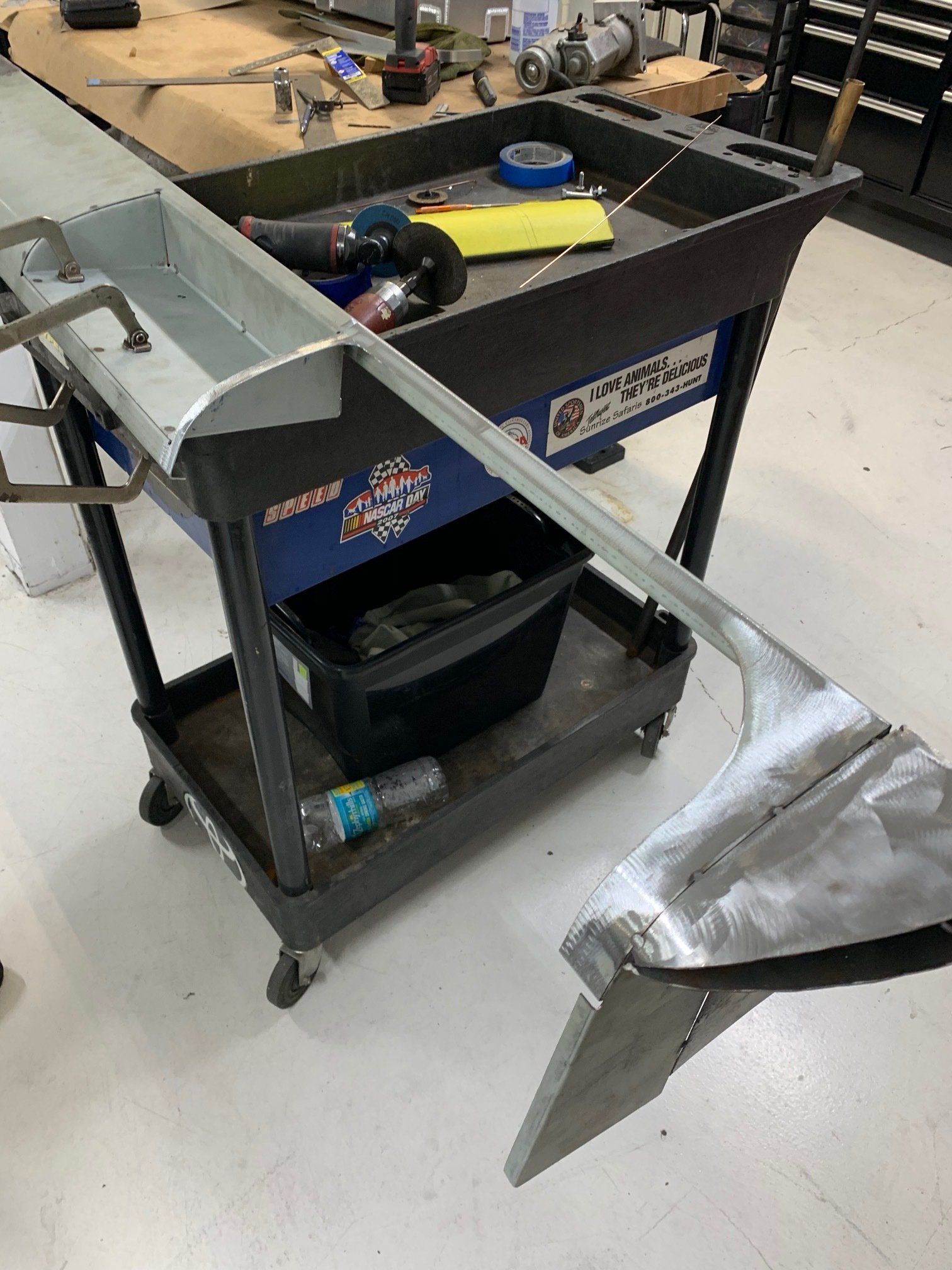Workshop cart with tools and supplies on shelves beside a metal part on a worktable