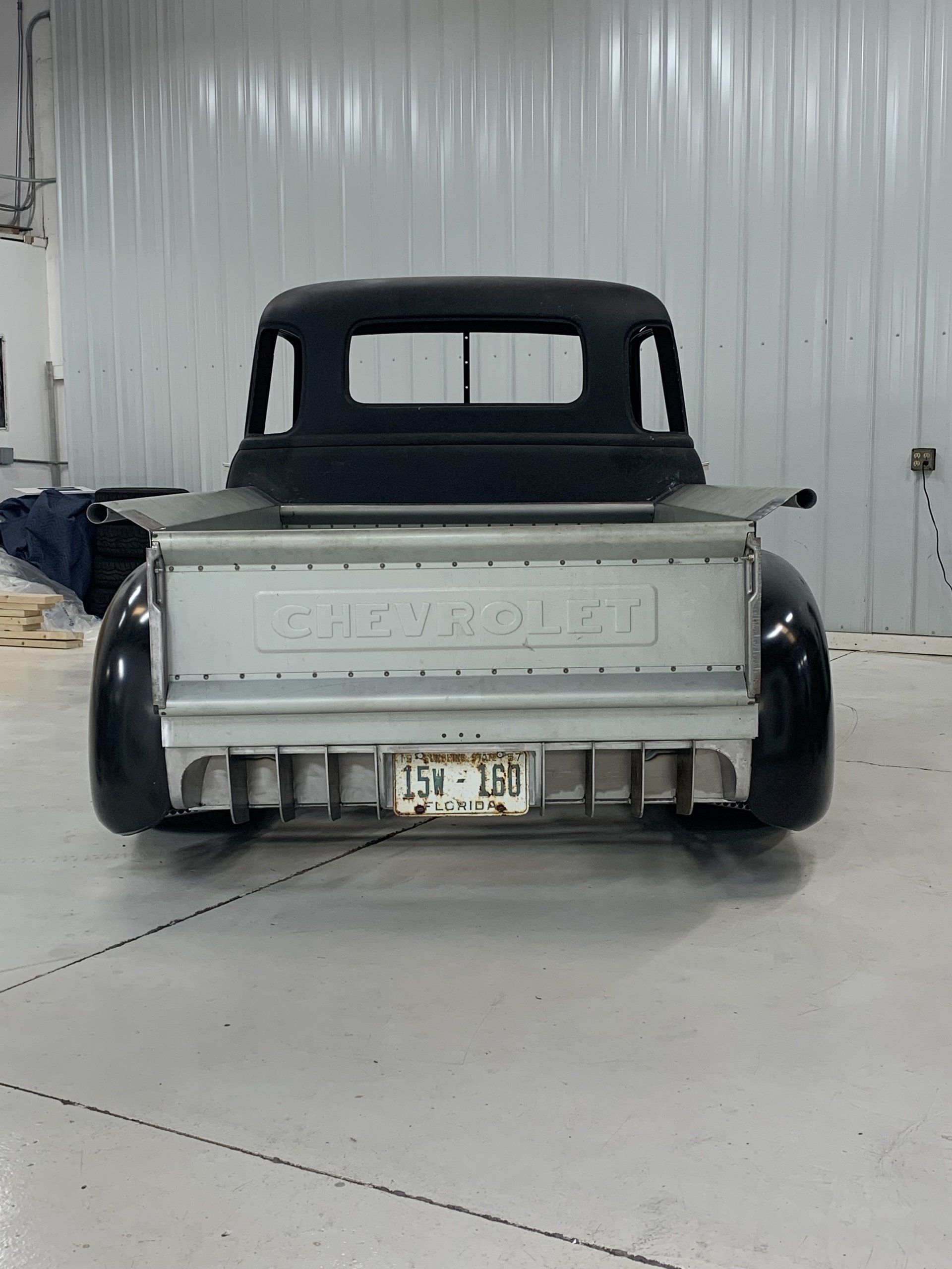 Rear view of a black vintage pickup truck in a garage, showing an unfinished silver tailgate and rear fenders