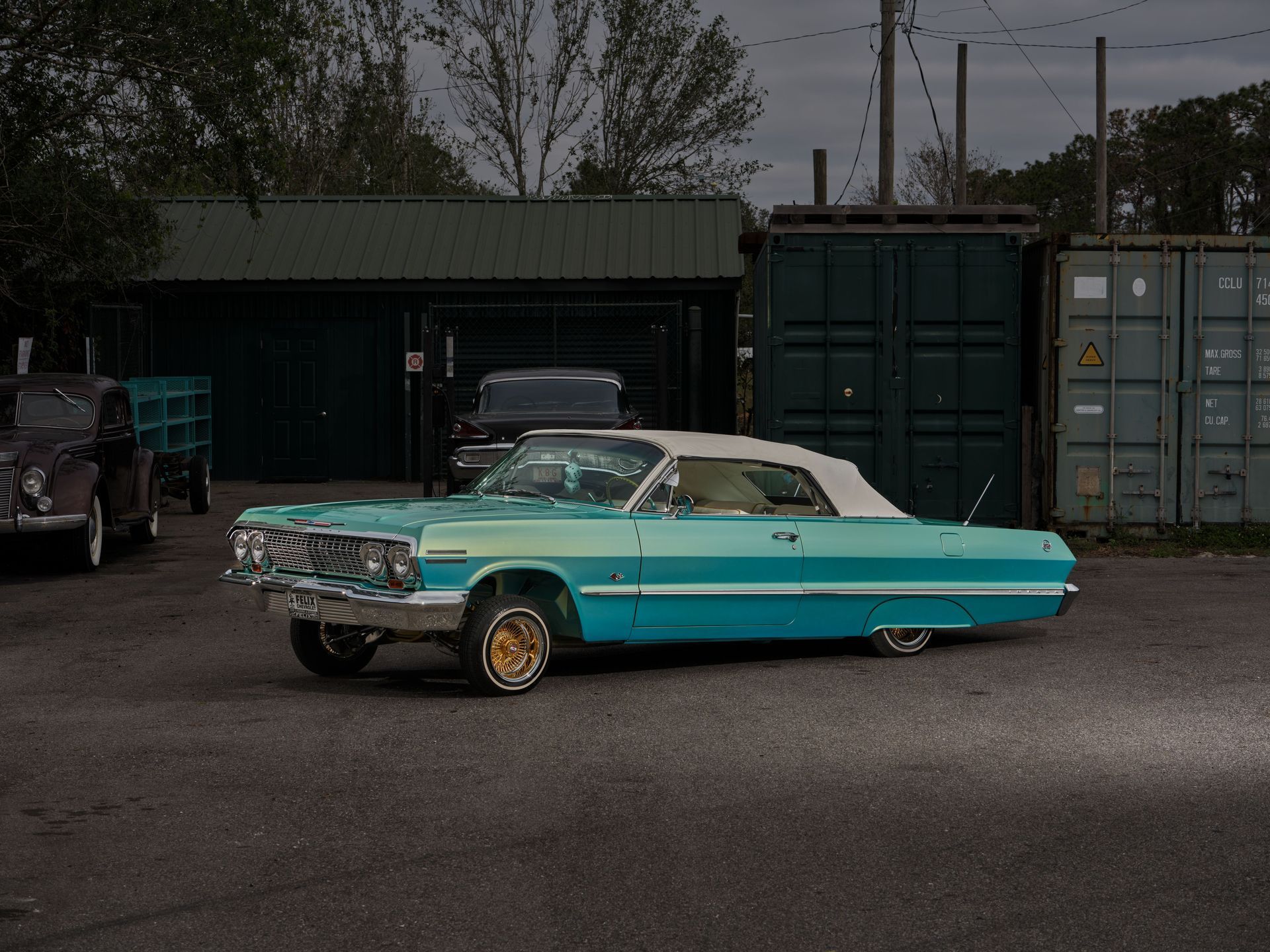 Turquoise classic convertible parked in an industrial lot beside shipping containers