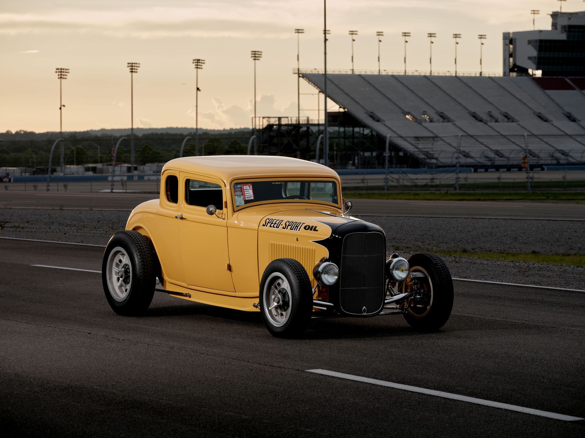 Yellow vintage hot rod on a racetrack with stadium bleachers in the background