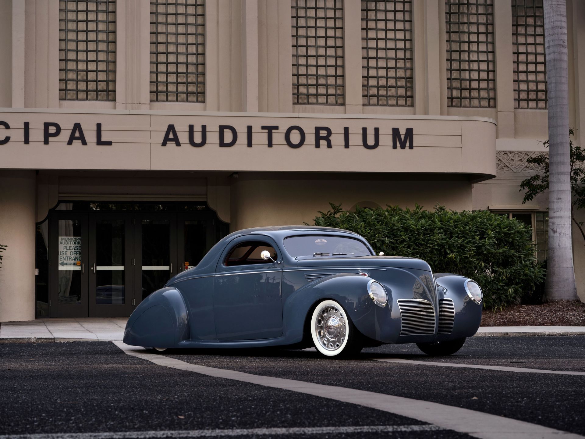 Blue vintage hot rod parked in front of a municipal auditorium.