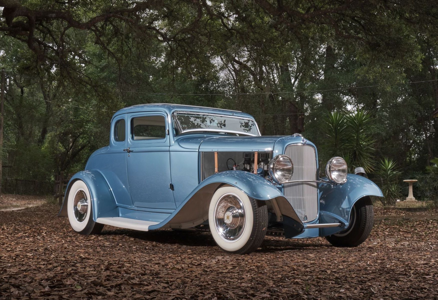 Vintage blue classic car parked on a leaf-covered road in a wooded setting
