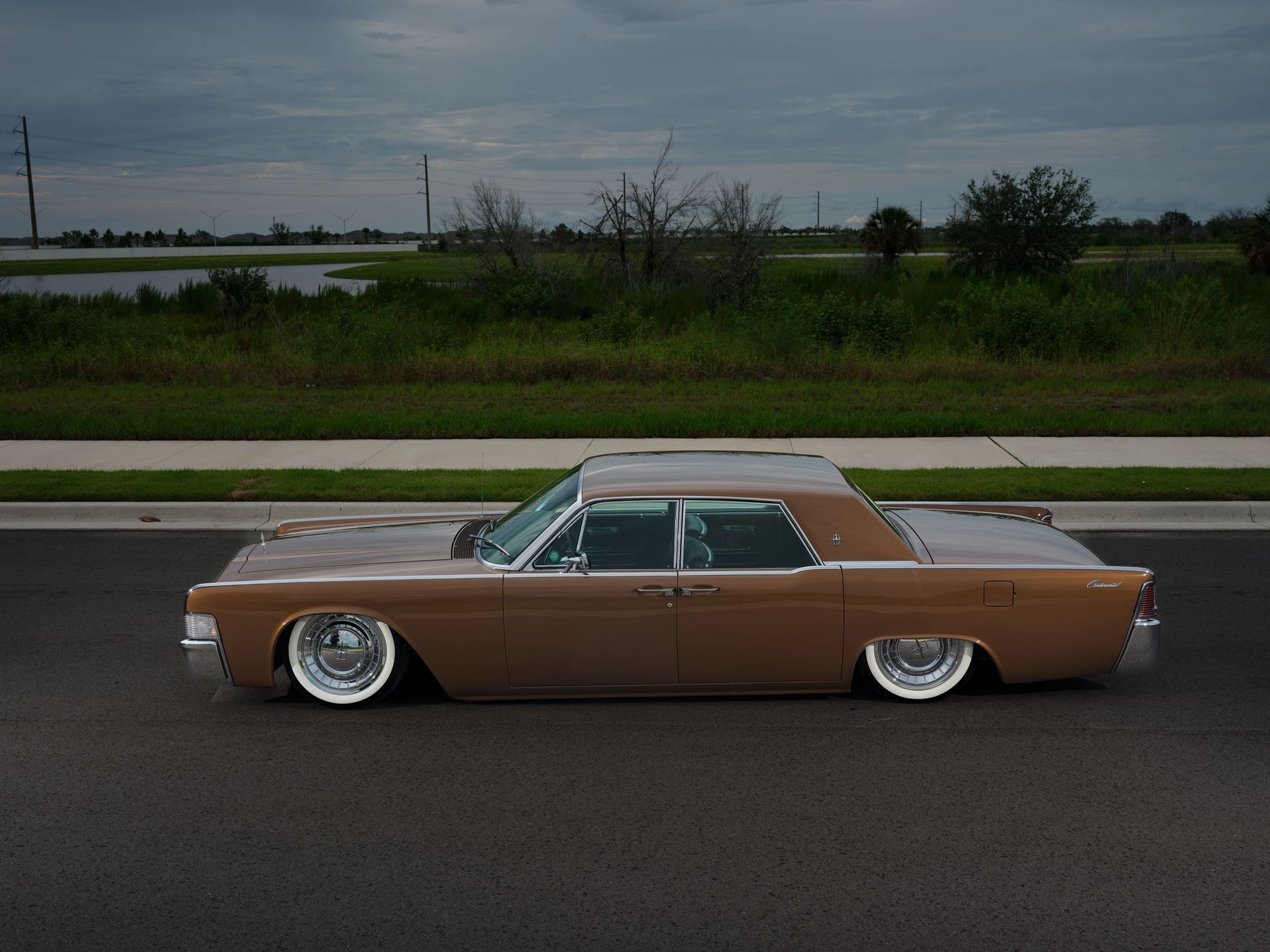 Lowered brown classic car parked on a road beside a grassy field under a cloudy sky