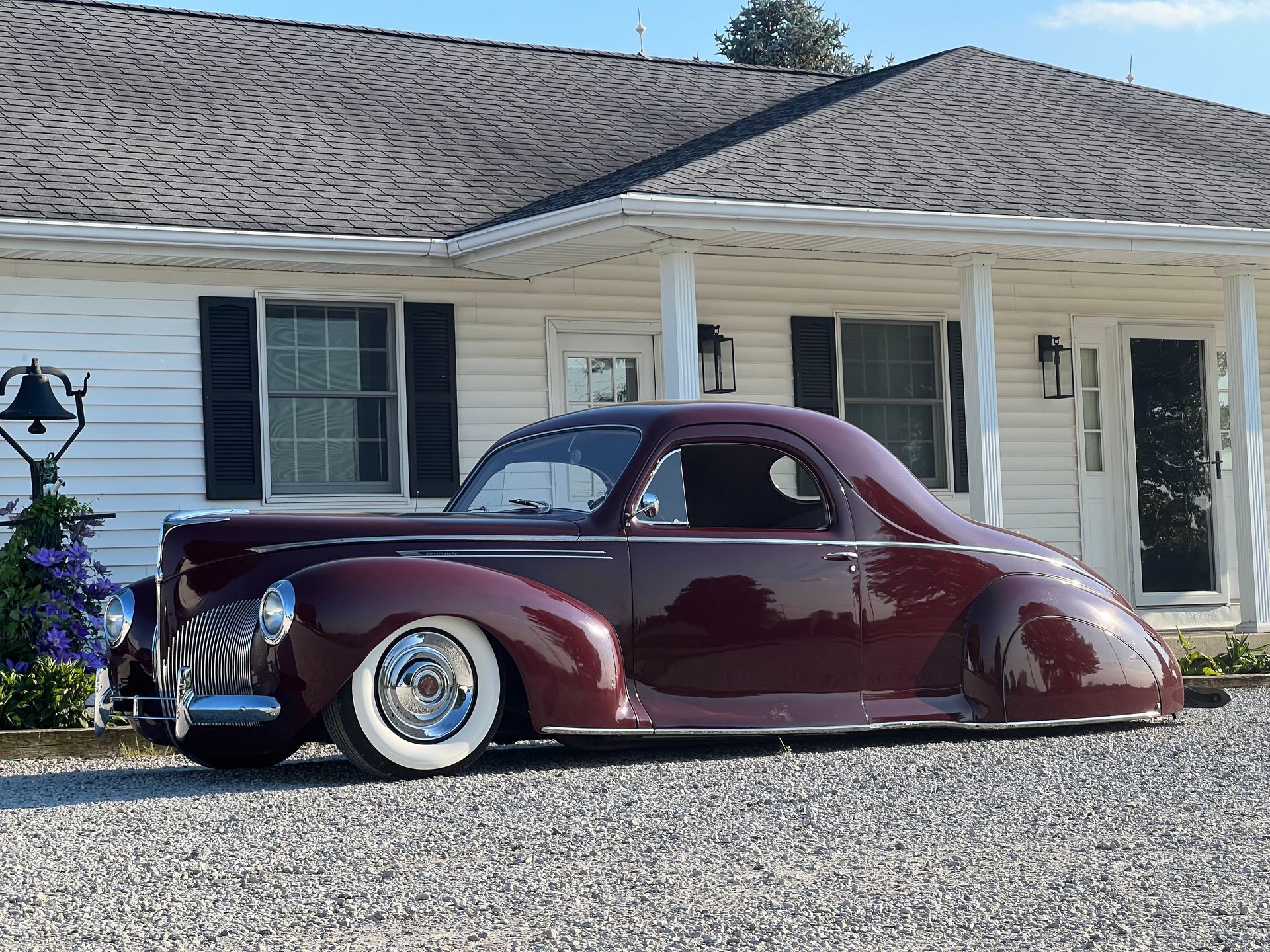 Classic burgundy hot rod parked in front of a white house with black shutters.