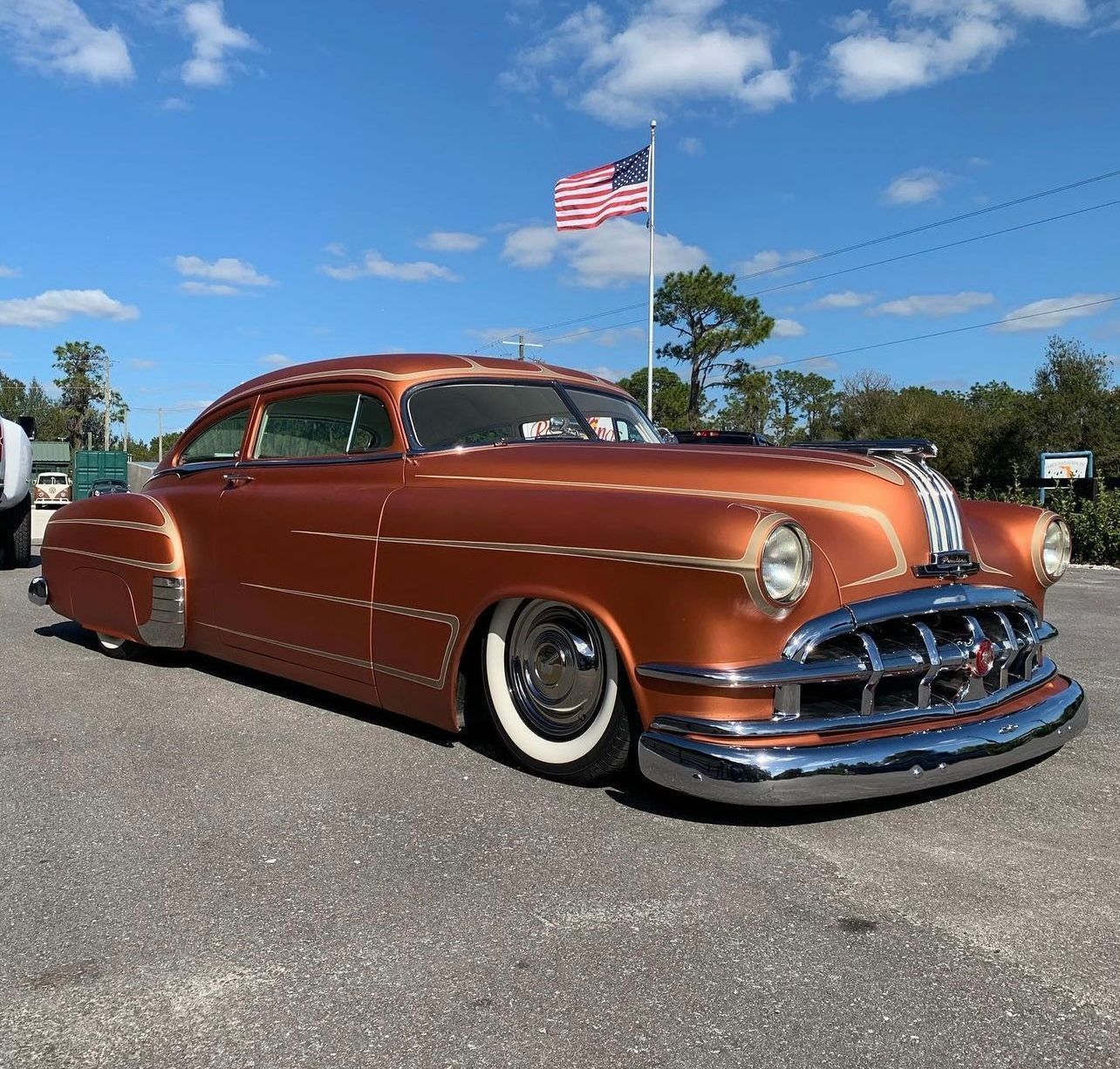 Custom copper lowrider car parked under a blue sky with an American flag waving behind it