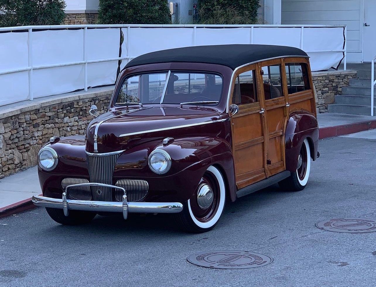 Vintage burgundy and wood-paneled station wagon parked on a street by a stone wall and white fence