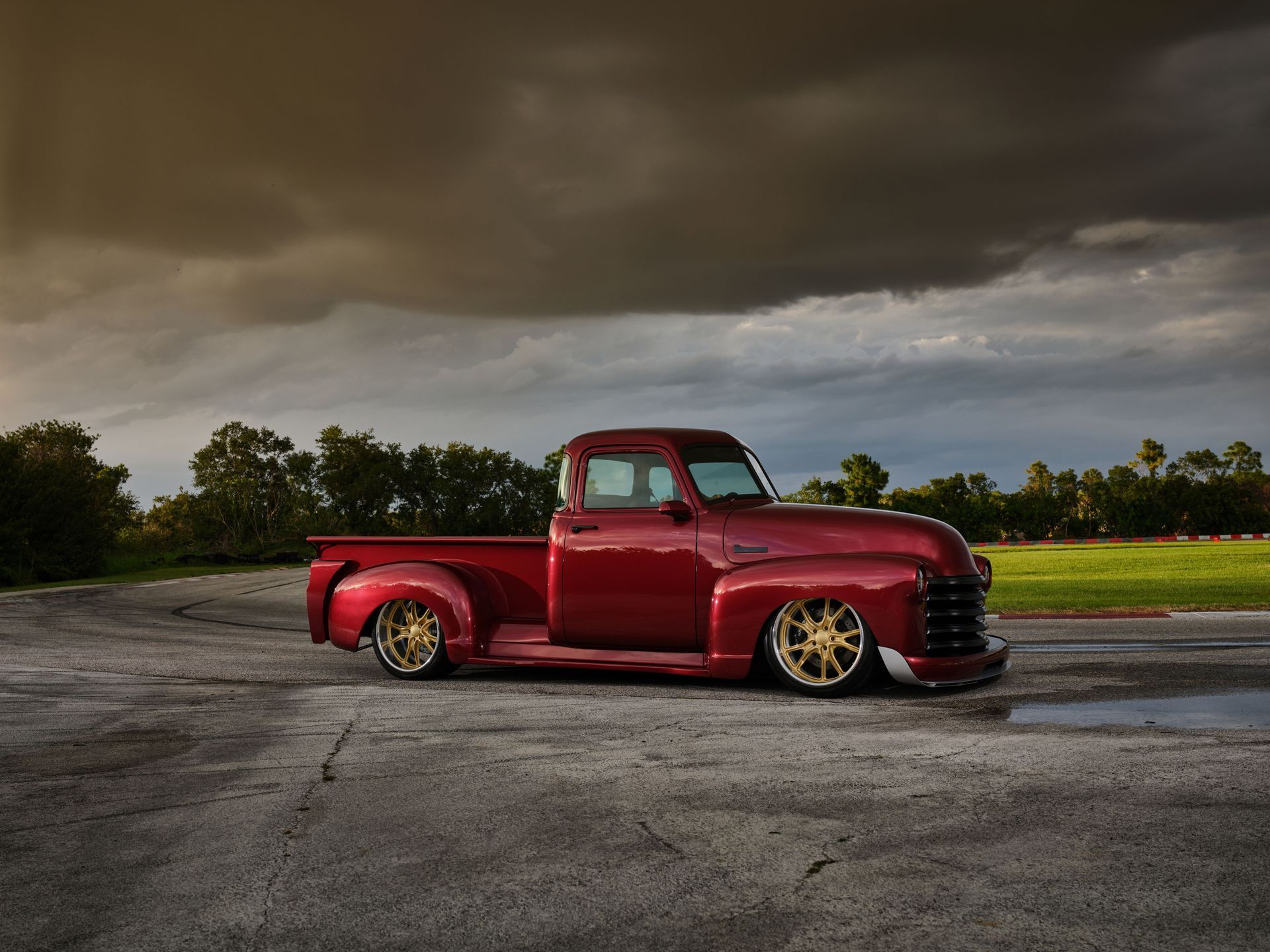Red vintage pickup truck parked on pavement under a cloudy sky in an open field.