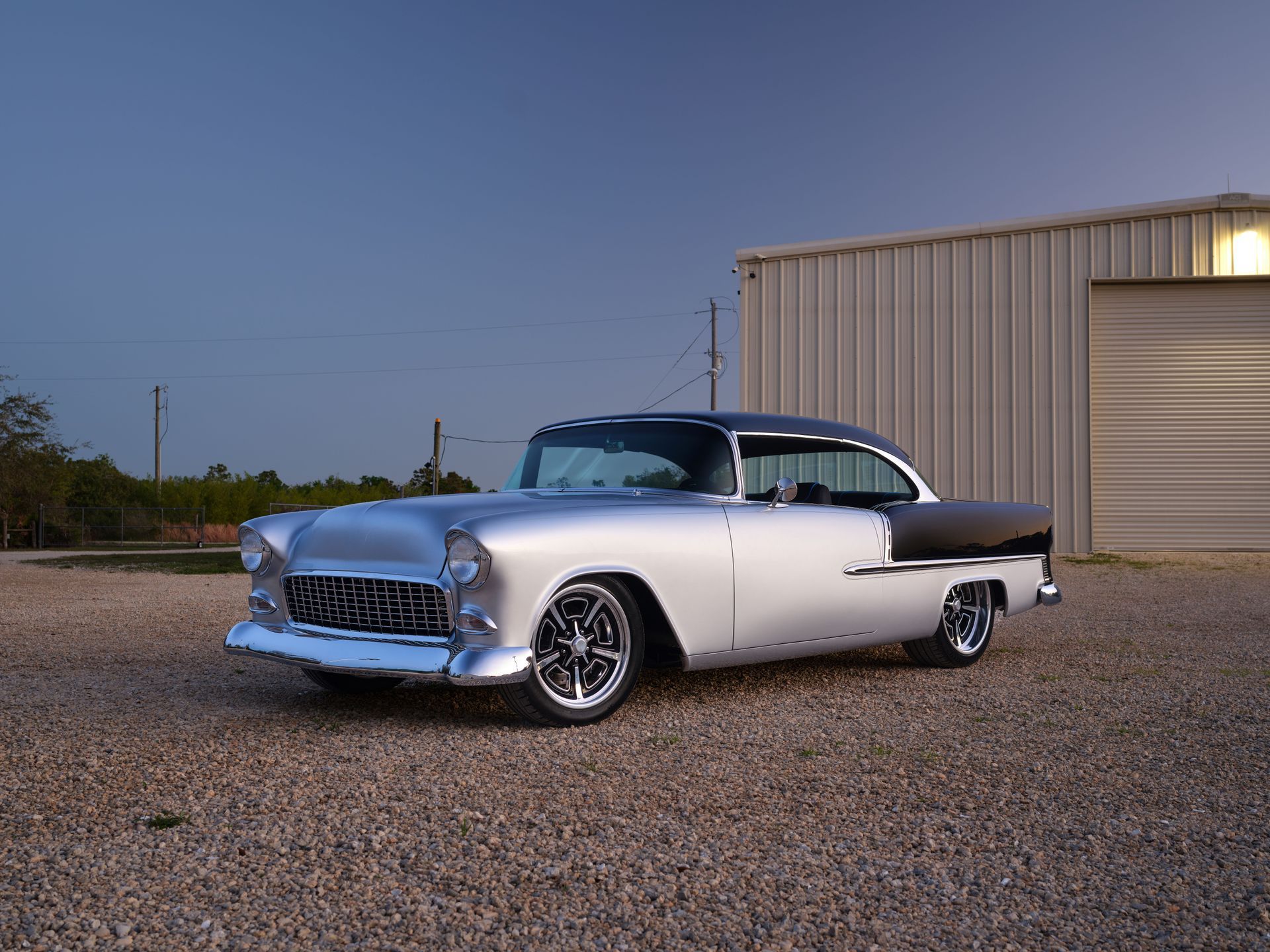 Classic silver-and-black vintage car parked on gravel beside a metal building at dusk