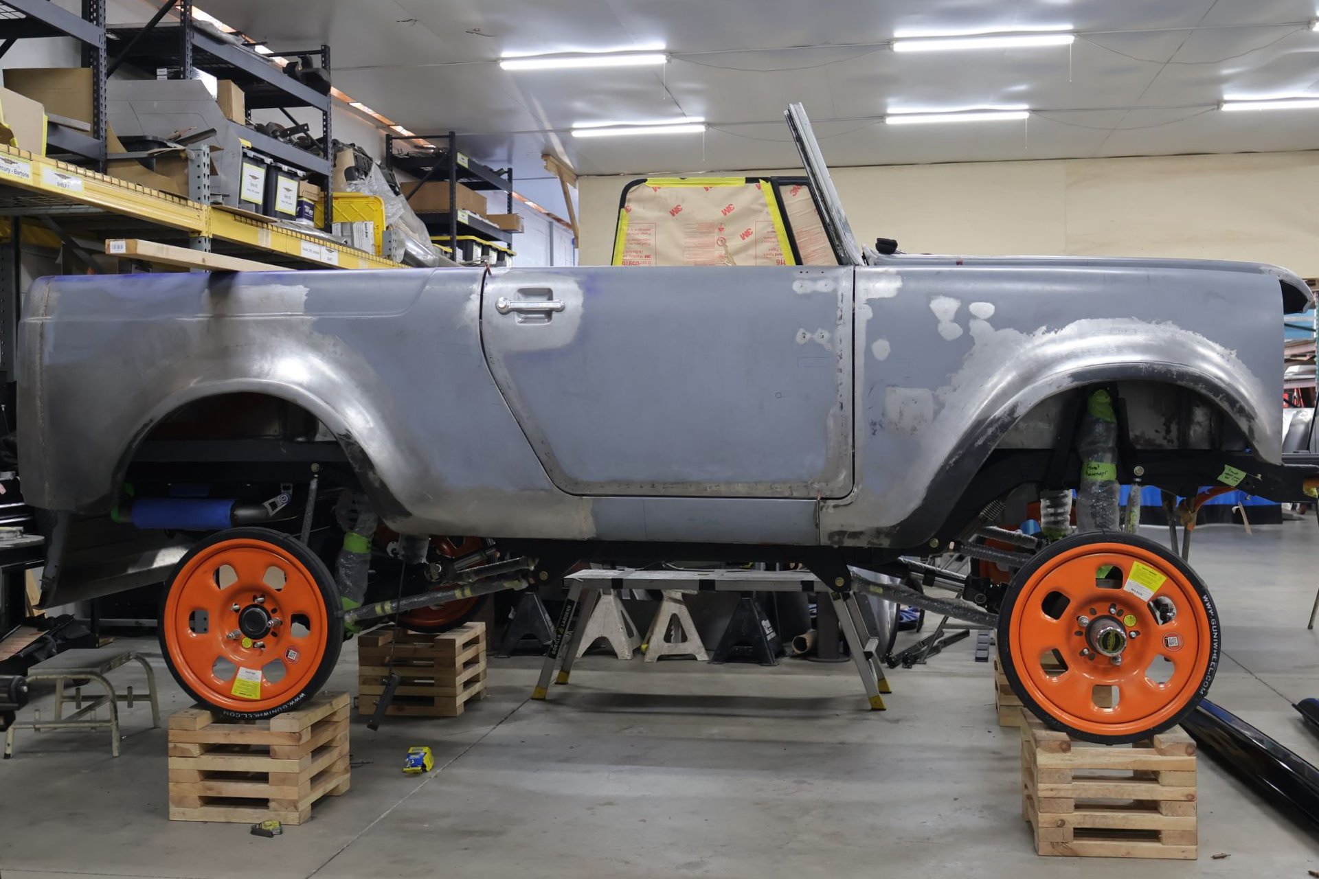 Gray classic car body on jack stands in a workshop, with orange wheels stacked on pallets nearby