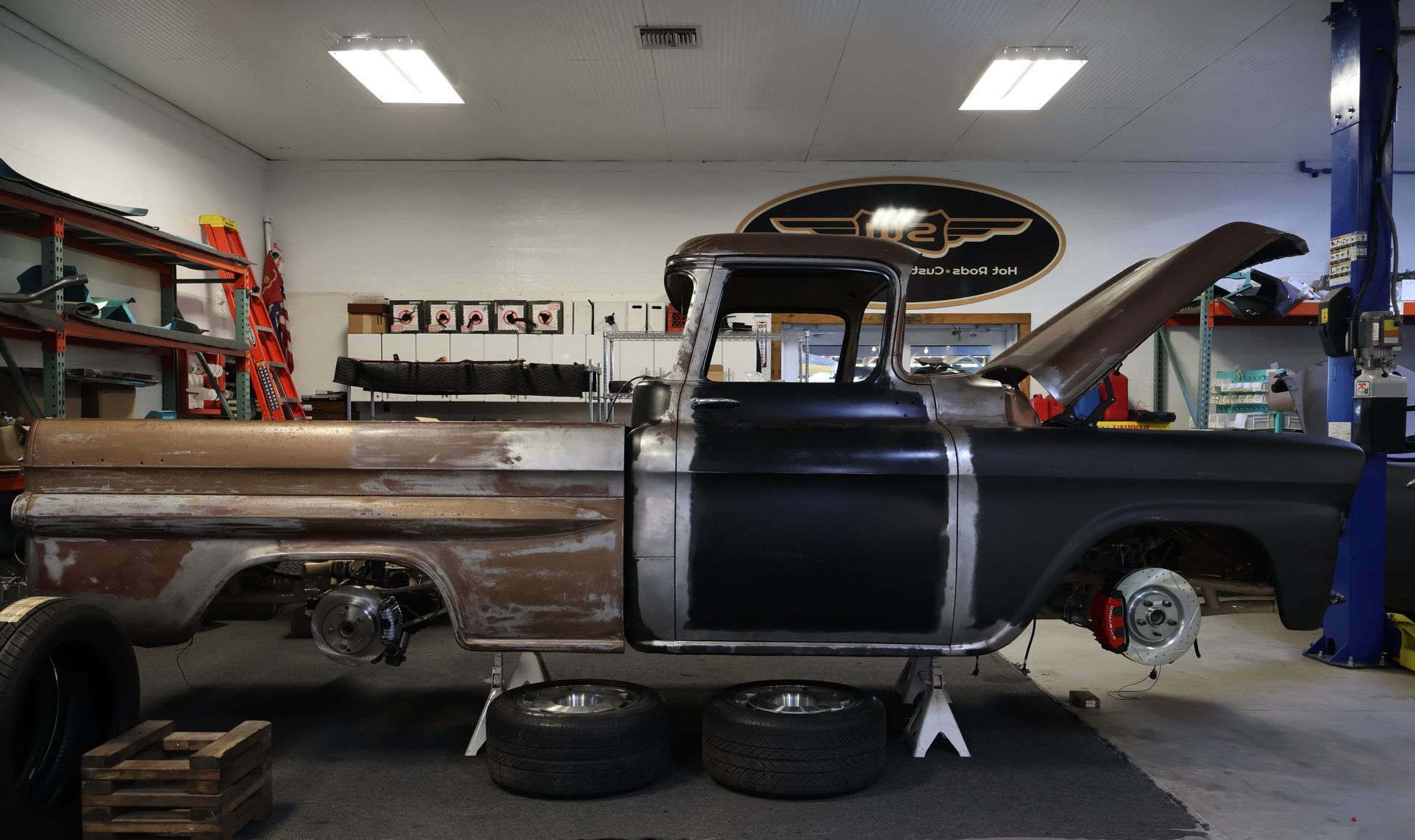 Classic rusty black-and-brown pickup truck in a garage, with the hood open and body partly restored.