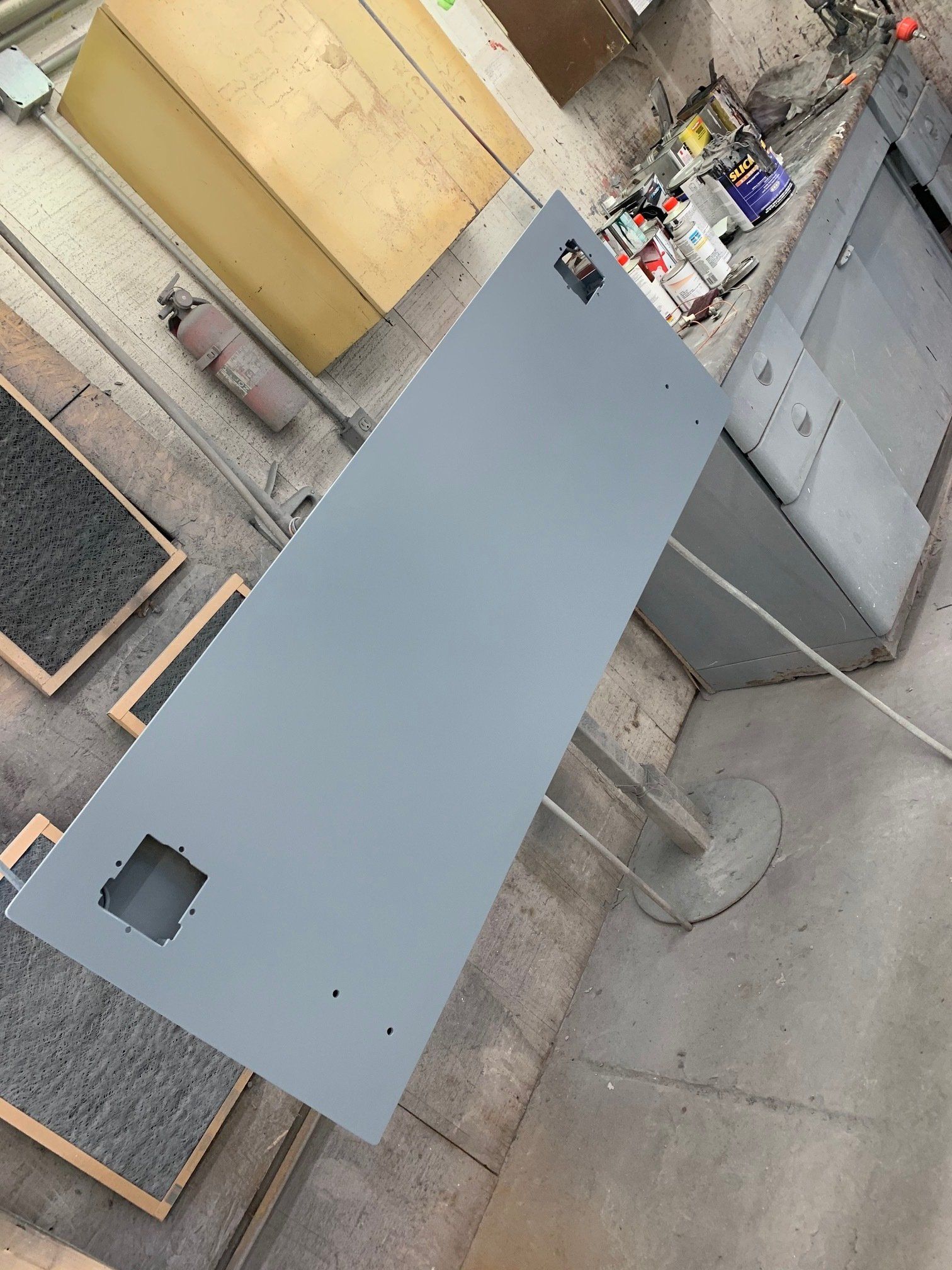 Gray metal panel laid on a workbench in an industrial workshop, with cutouts and tools nearby.