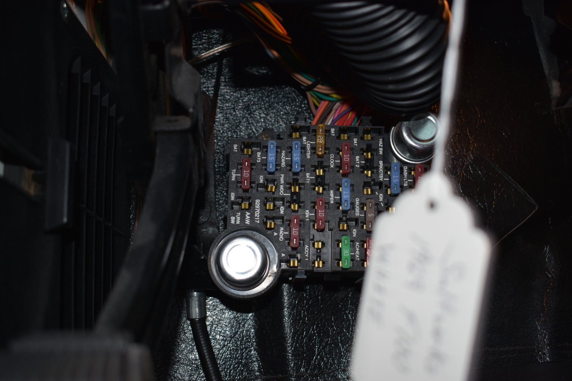 Close-up of a car fuse box with colorful fuses and a white hanging tag in a dark compartment