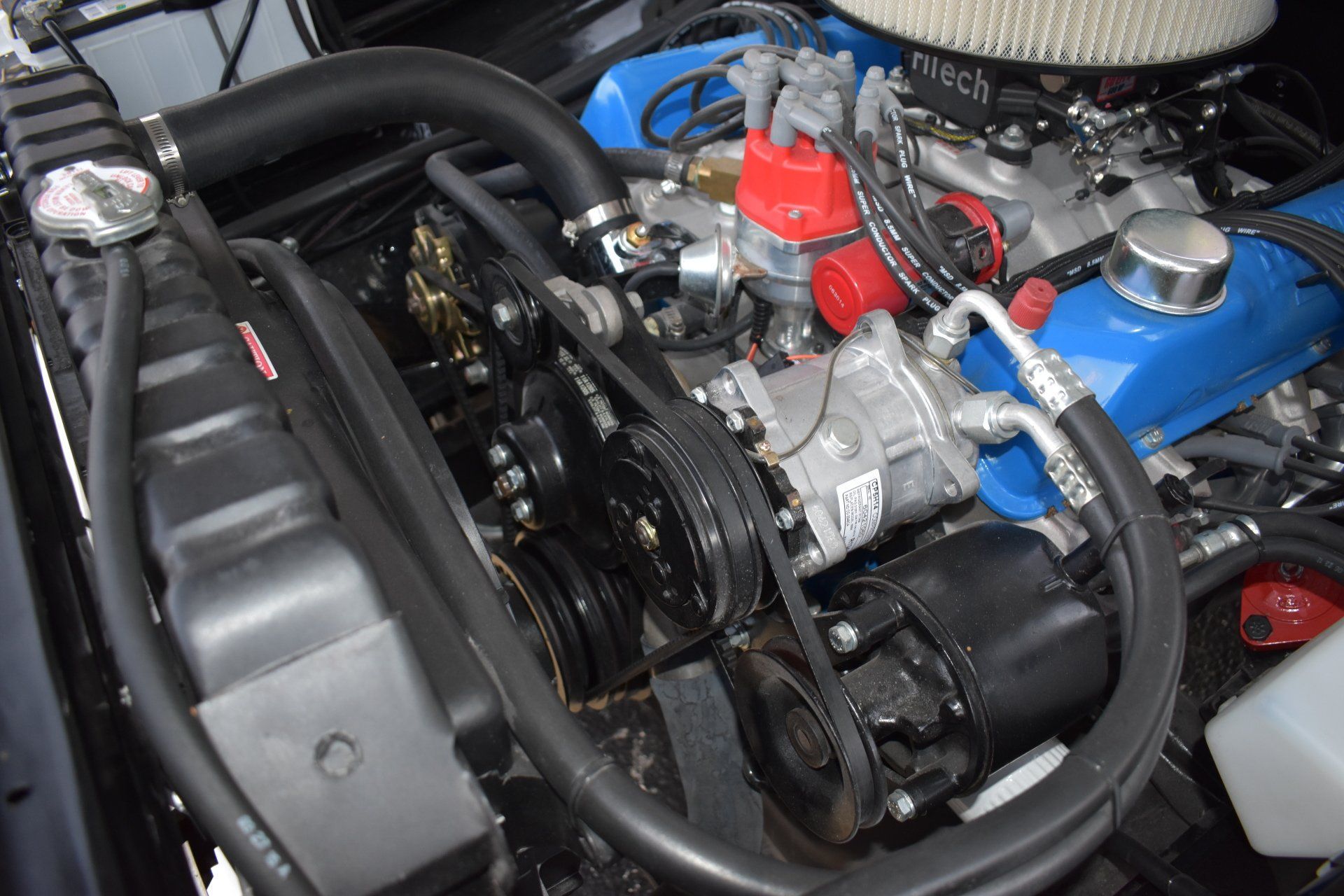 Close-up of a classic car engine with blue valve covers, black belts, and chrome carburetor components.