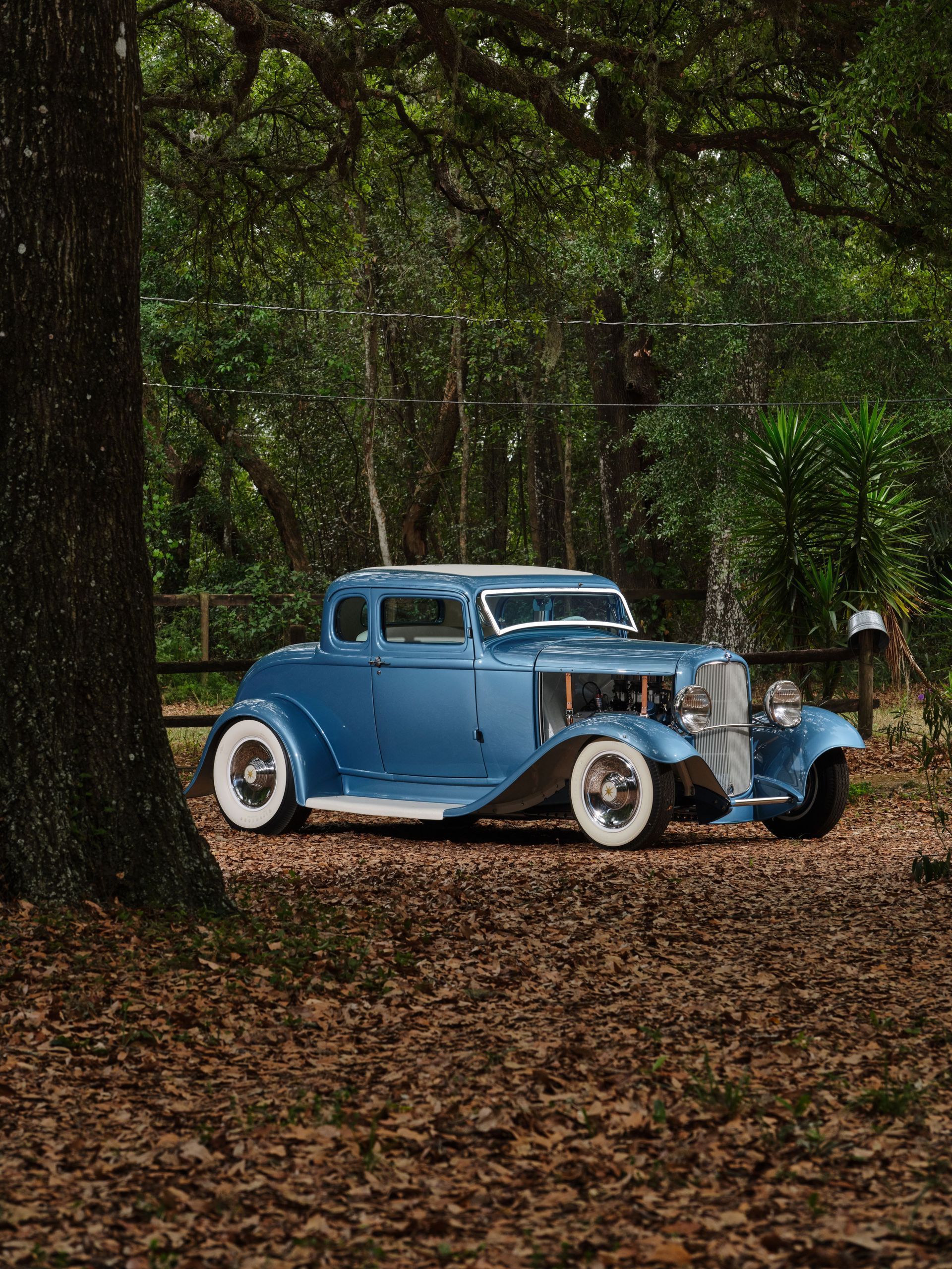 Blue vintage car parked on a leaf-covered path in a wooded forest clearing