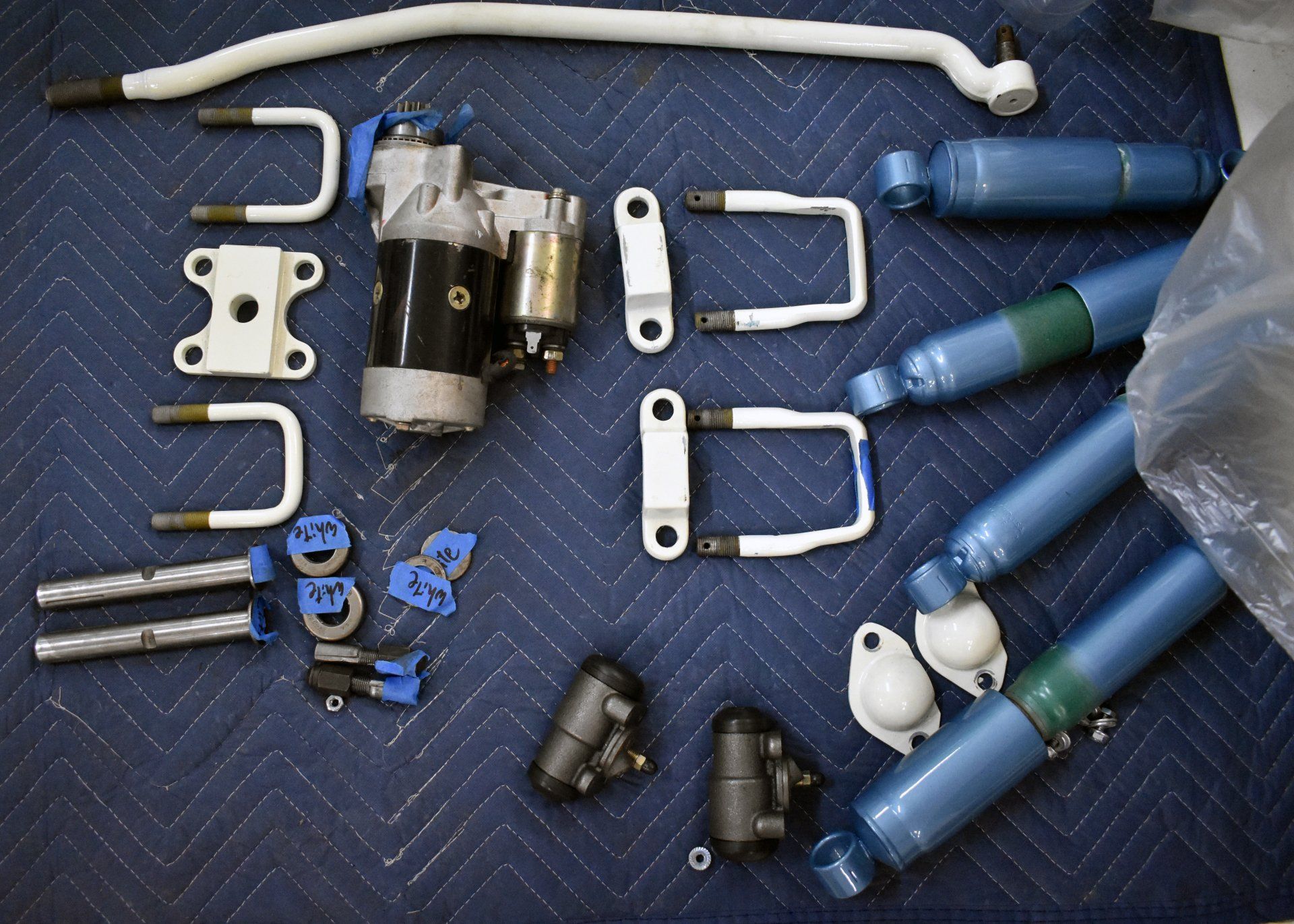 Assorted metal and blue automotive parts laid out on a blue mat, including rods, brackets, and motor pieces
