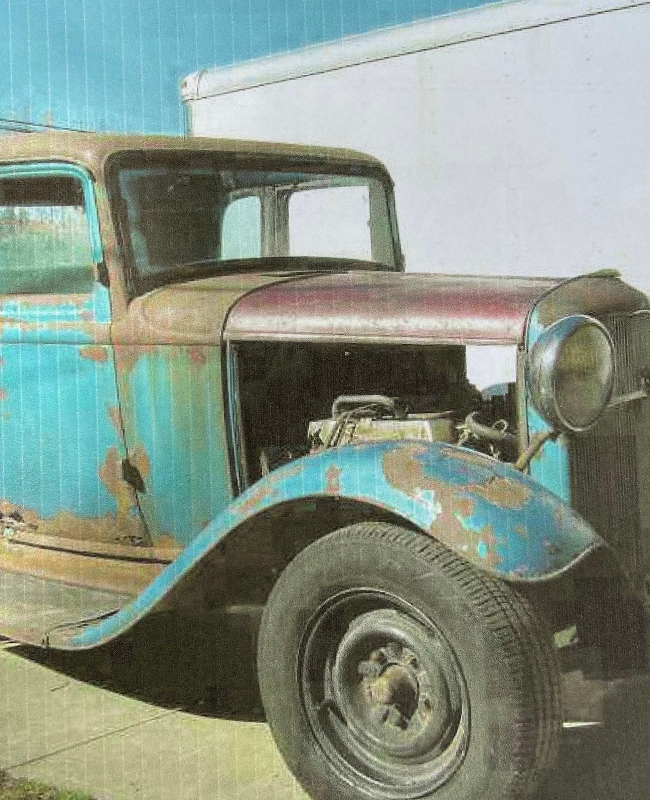 Rusty turquoise vintage car with black fender and large headlight, parked against a plain wall.