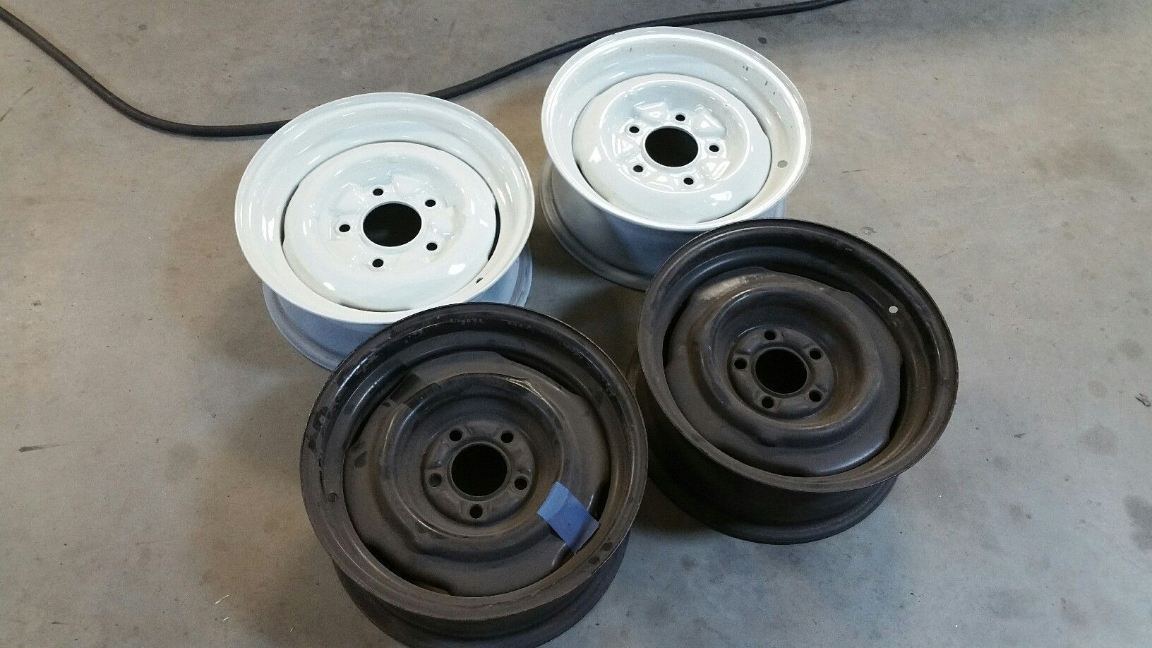 Four vehicle wheels/rims, two white and two black, stacked on a concrete floor.