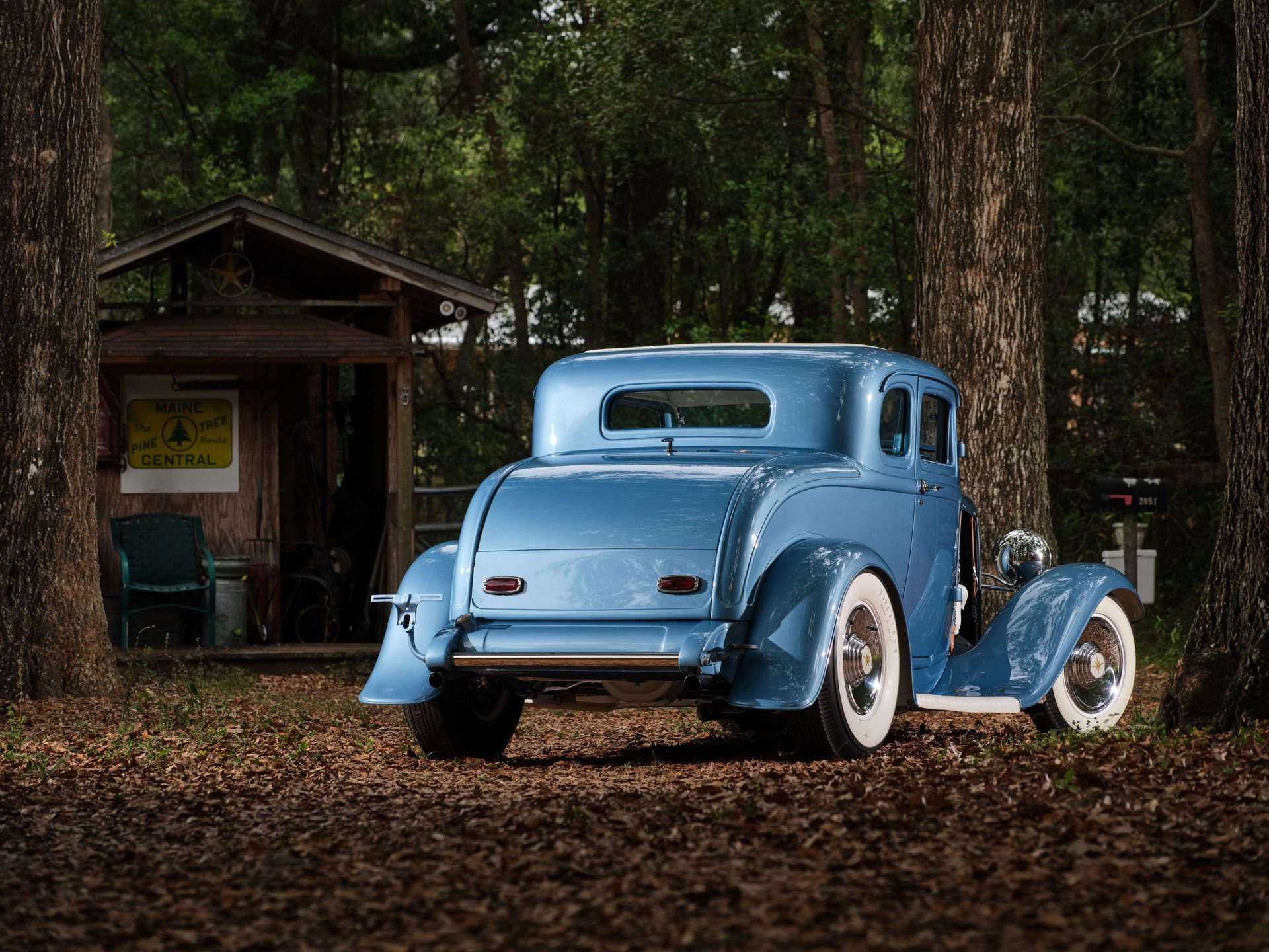 Light blue vintage car parked on leaf-covered ground beside a small wooden cabin in a forest