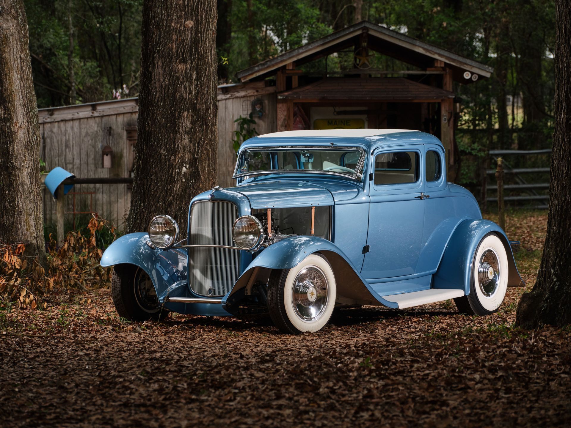 Light blue vintage car parked in a wooded area beside a small cabin.